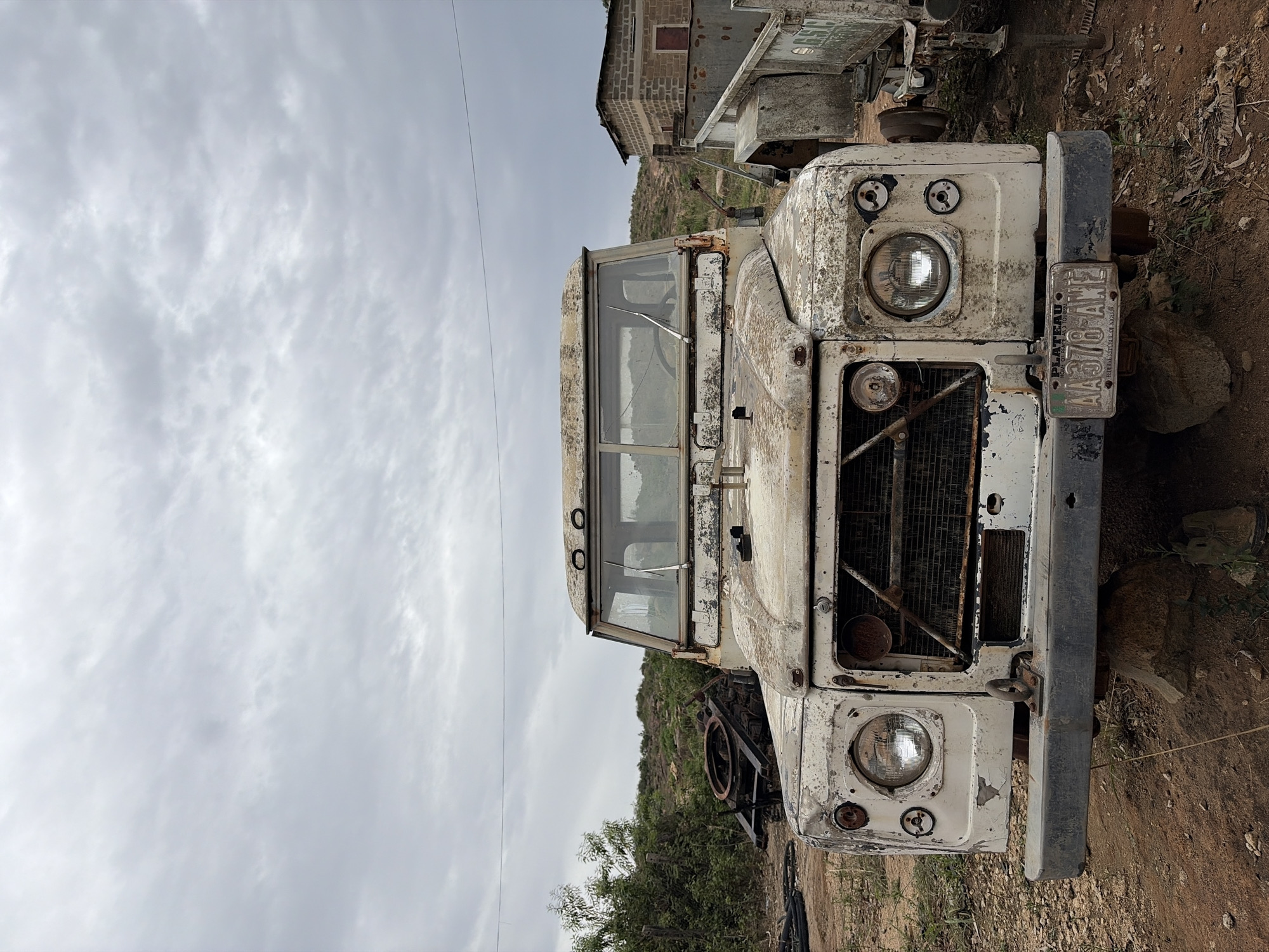 The image shows a dilapidated, white Land Rover vehicle with the license plate "AA378 AME," seemingly abandoned in an outdoor environment in Gura Top, Nigeria. The vehicle's front is heavily weathered and marked with rust. Its headlamps are visible, along with a corroded radiator grill and a partial view of the windshield. A building and some vegetation are partially visible in the background. The ground is dry and covered with dirt and sparse plant life. The sky is overcast.