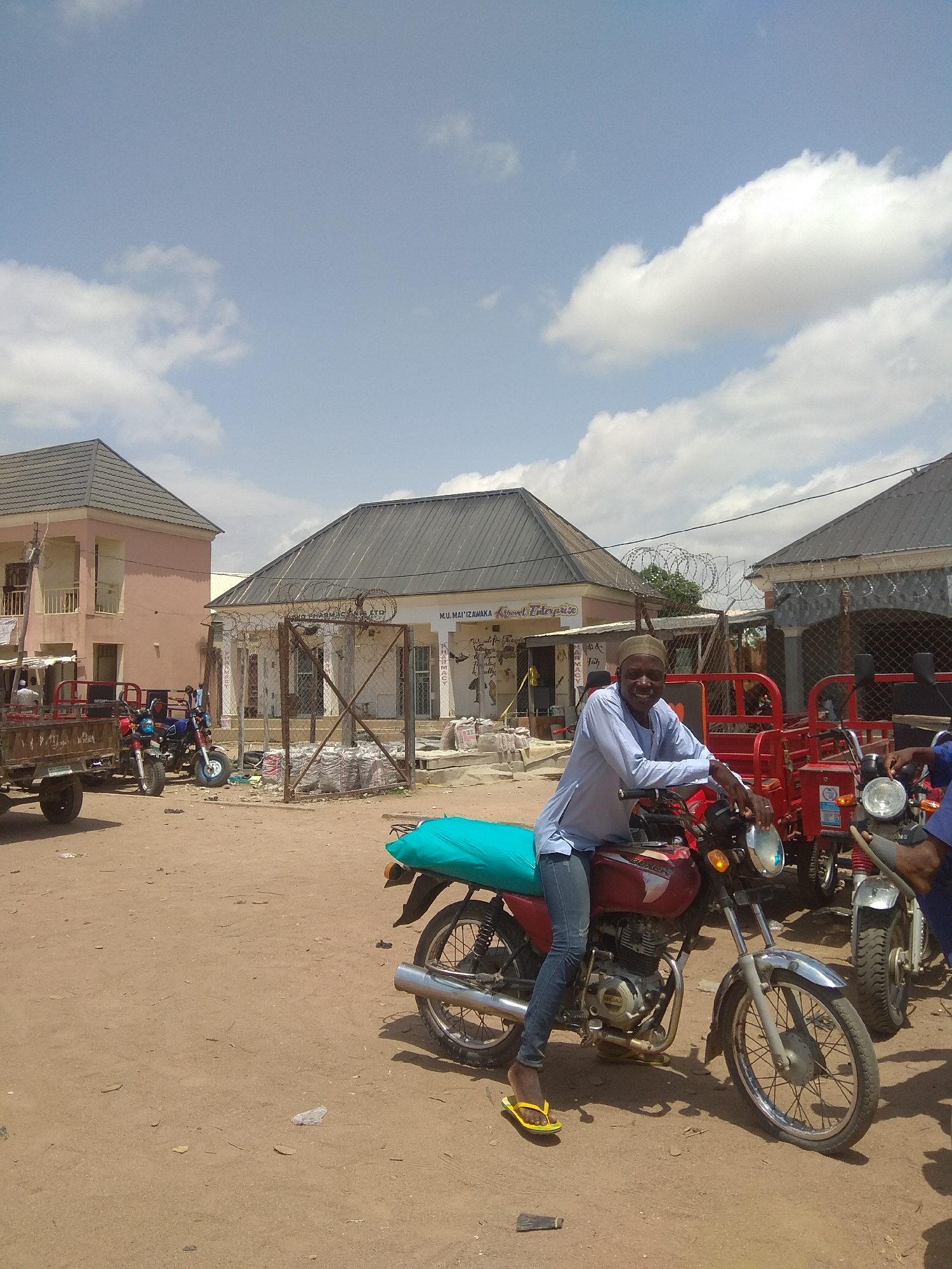 This media file shows an outdoor scene in Jalingo, Nigeria, under a bright, cloudy sky. The central subject is a man wearing a light blue traditional outfit and a skullcap, seated on a red motorcycle. He is smiling and looking towards the camera, appearing relaxed and cheerful. The motorcycle has a turquoise tarp draped over its rear.
In the background, there are several buildings. One is a two-story pink structure with a balcony, and another appears to be a commercial establishment with signage that is partially visible. There is also a building with a dark, sloped roof and wire fencing in front of it, suggesting a commercial or administrative area.
Several other vehicles are present, including another motorcycle, a red motorized tricycle, and a dark-colored trailer attached to a vehicle on the left side of the frame. Bags of goods are stacked near one of the buildings. The ground is covered in dirt and sand, typical of a dusty environment. The presence of multiple motorcycles and a trailer suggests a marketplace or a transportation hub. The overall impression is of a typical day in a bustling Nigerian town, with people going about their business. The time of day appears to be late morning or early afternoon, given the strong sunlight and shadows.