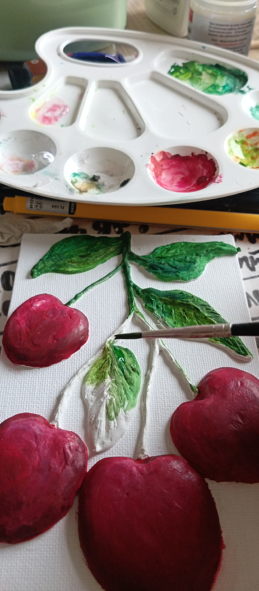 The image shows a close-up view of an art project in progress. The subject is a painting of cherries on a white textured canvas. Three plump, red cherries are visible, along with green leaves and stems. A paintbrush is actively applying green paint to one of the leaves, which is partially painted white. Above the painting, a white plastic artist's palette displays a range of colors, including various shades of red and green. A yellow marker rests between the painting and the palette. The artwork appears to be still life, focusing on the textures and colors of the cherries and leaves. The location is Unknown, Unknown.