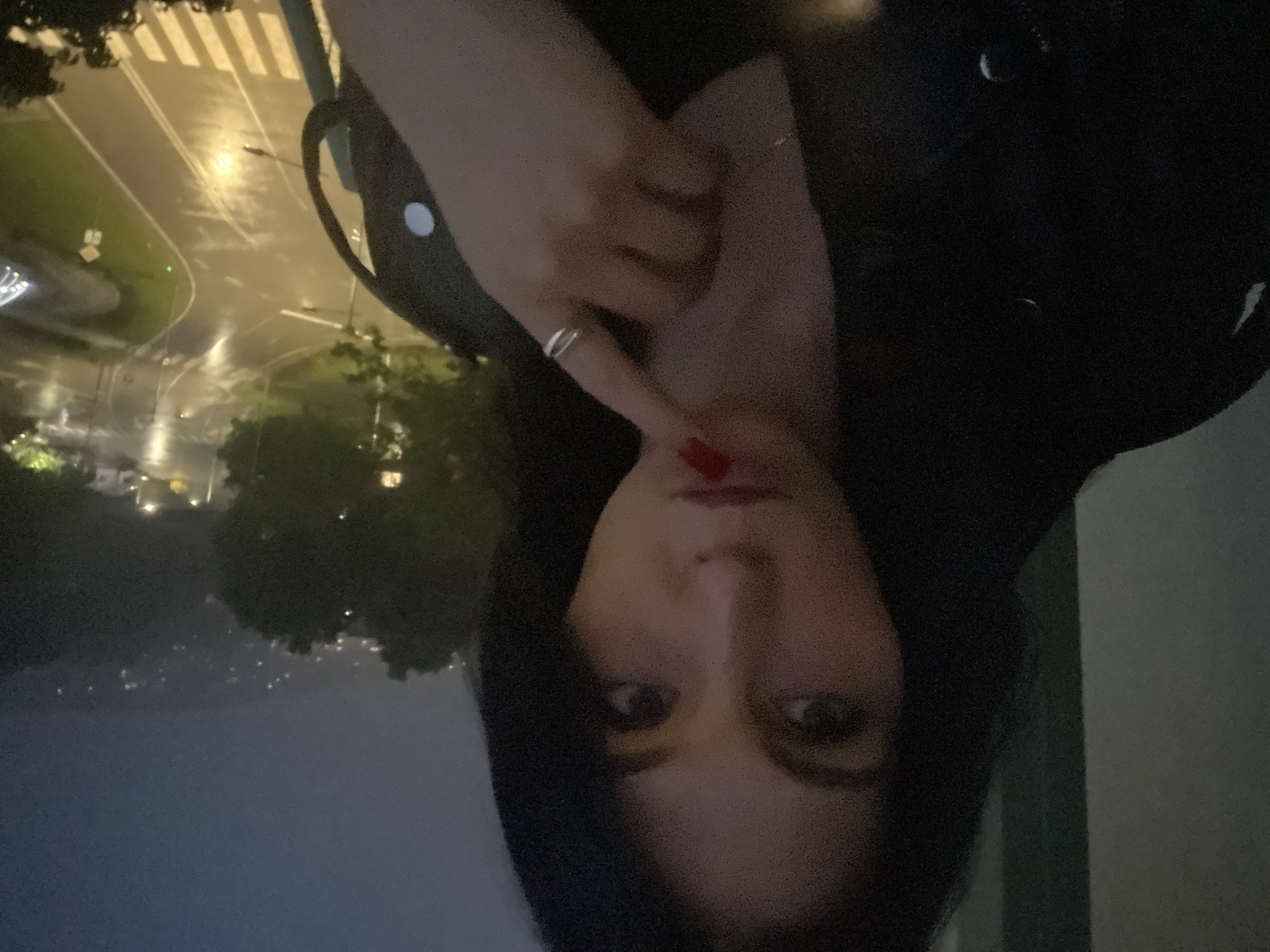 The image depicts a person, likely a woman, seen from the chest up, with a background of an urban or suburban night scene. The primary subject is looking directly at the viewer. Her dark hair frames her face, and her eyes are open. Her lips are partly visible and appear to have red lipstick. One hand is raised towards her face, with fingers slightly parted; red nail polish is visible on one fingernail, and a silver ring is on another finger. She appears to be wearing a dark garment, possibly a jacket or top.
The background, blurred and dark, shows an outdoor setting at night or dusk. A wet road, possibly from rain, is illuminated by streetlights, with distinct zebra crossings visible. Green areas with trees are adjacent to the road. In the far distance, faint lights suggest a city or residential area. The perspective indicates the photo was taken from an elevated position, possibly from a building or balcony overlooking the road and landscape. There is no visible text in the image.