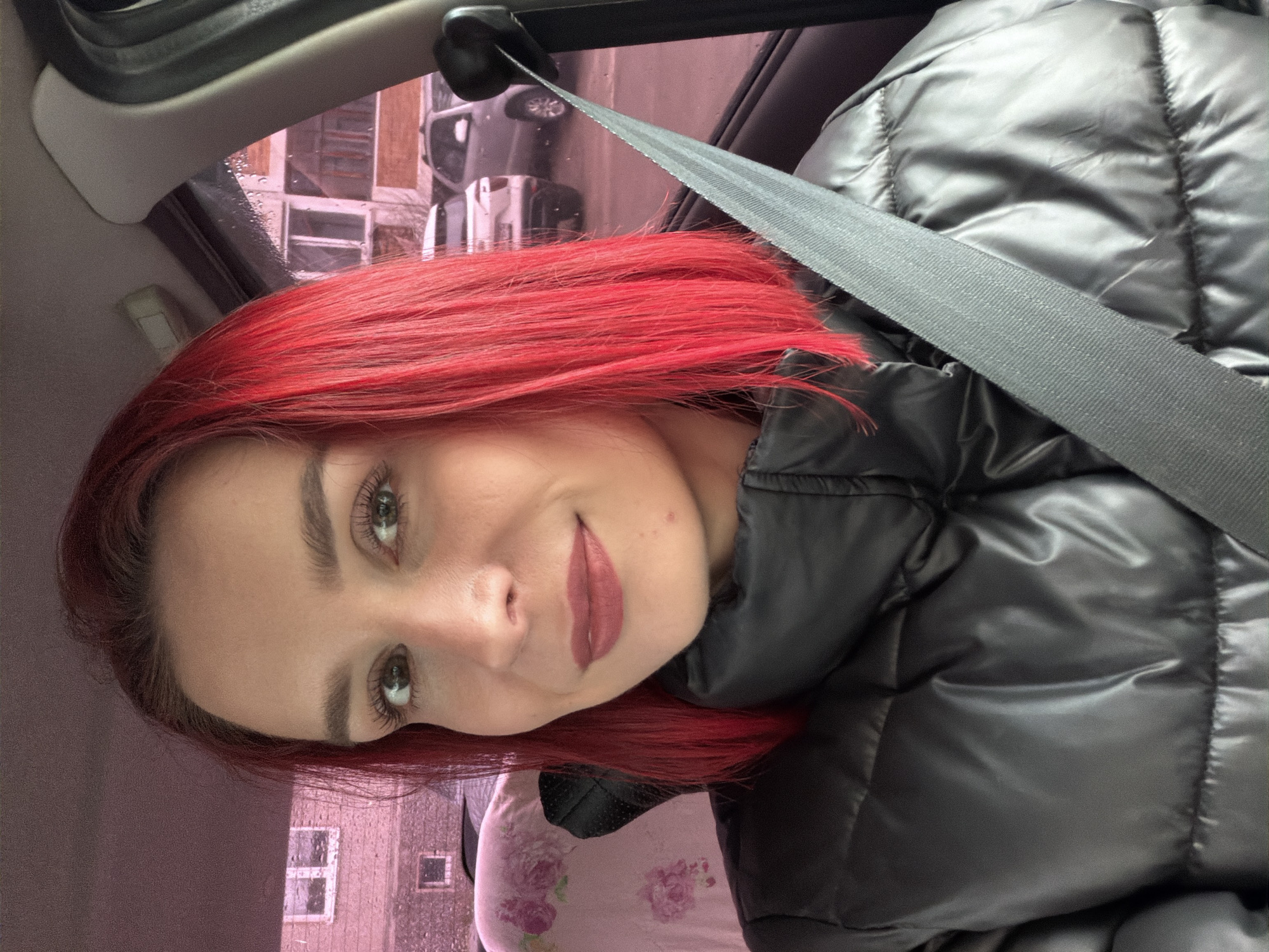 A woman with bright red hair is in a car wearing a black puffer jacket and a seatbelt. The background shows buildings through a rain-streaked window. The city is Chaikovsky, Russia.