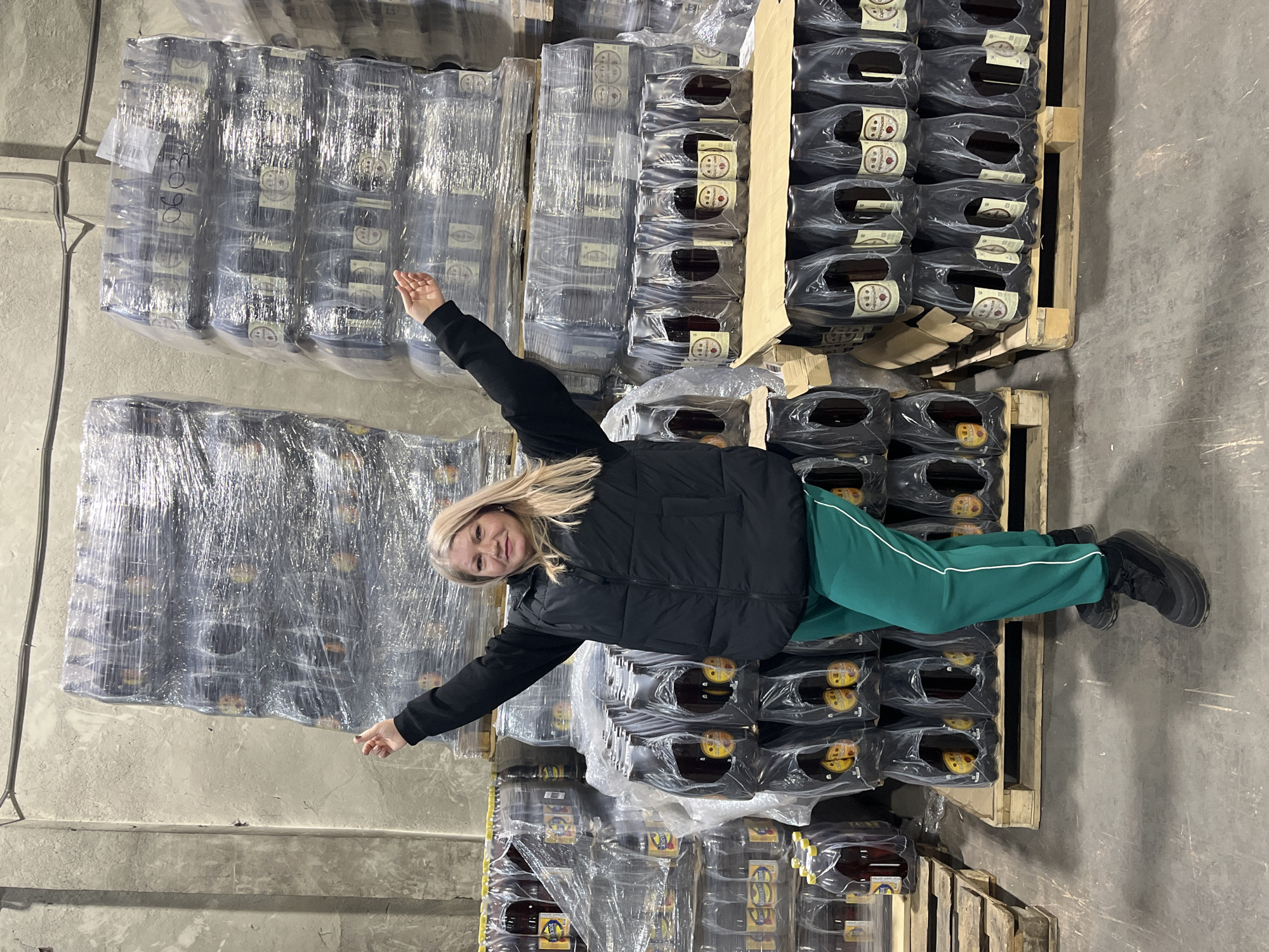 The image depicts an adult female standing within a warehouse or storage facility in Izhevsk, Russia. This location is identified by the presence of numerous stacks of "САБЕЛЬНОЕ" (Sabelnoye) branded kvass bottles, a product associated with a production company in Izhevsk.
The female, positioned centrally and facing the viewer, has shoulder-length blonde hair. She wears a black puffer vest over a black long-sleeved garment, green track pants with a white stripe down each outer leg, and black athletic shoes. Her arms are raised above her head, bent at the elbows, with her palms open and fingers slightly splayed. A faint smile is visible on her face.
She is surrounded by tall stacks of dark-colored plastic beverage bottles, predominantly "САБЕЛЬНОЕ" kvass, arranged in shrink-wrapped bundles on wooden pallets. To her left, additional stacks include smaller, clear plastic bottles labeled "ЖИТЕЙСКОЕ КВАС" and "МЕДОВУХА." On the clear shrink-wrap of one upper pallet stack, the hand-written alphanumeric sequence "06.03" is visible. The floor is dark grey concrete, and the background consists of rough, unpainted concrete walls and vertical concrete pillars. Dark utility cables are attached to the upper section of the rear wall.