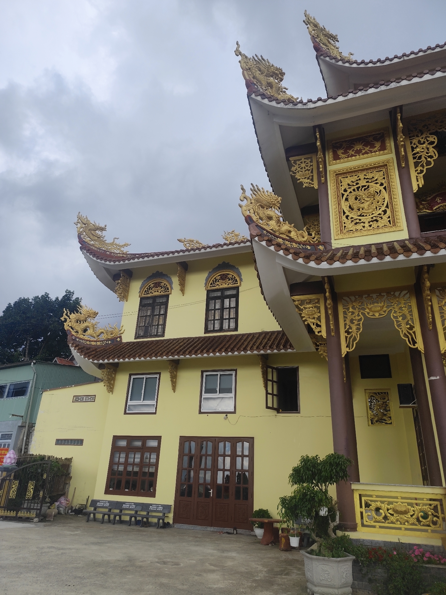 The image depicts a bright yellow building with ornate, gold dragon roof decorations, a common feature in Vietnamese temples. The building has multiple stories, with a prominent, curved roofline adorned with detailed dragon motifs. There are several windows, some with dark brown frames and others with white frames. On the ground floor, there are large brown wooden doors. In front of the building, a concrete courtyard is visible, with a row of stone benches. To the left, a black metal fence encloses some greenery and decorative elements, including what appears to be a pink flower display. A small tree in a decorative pot sits to the right of the main building entrance. The sky above is overcast with grey clouds. The context of Xuân Thành, Vietnam, suggests this is likely a temple or a cultural building.