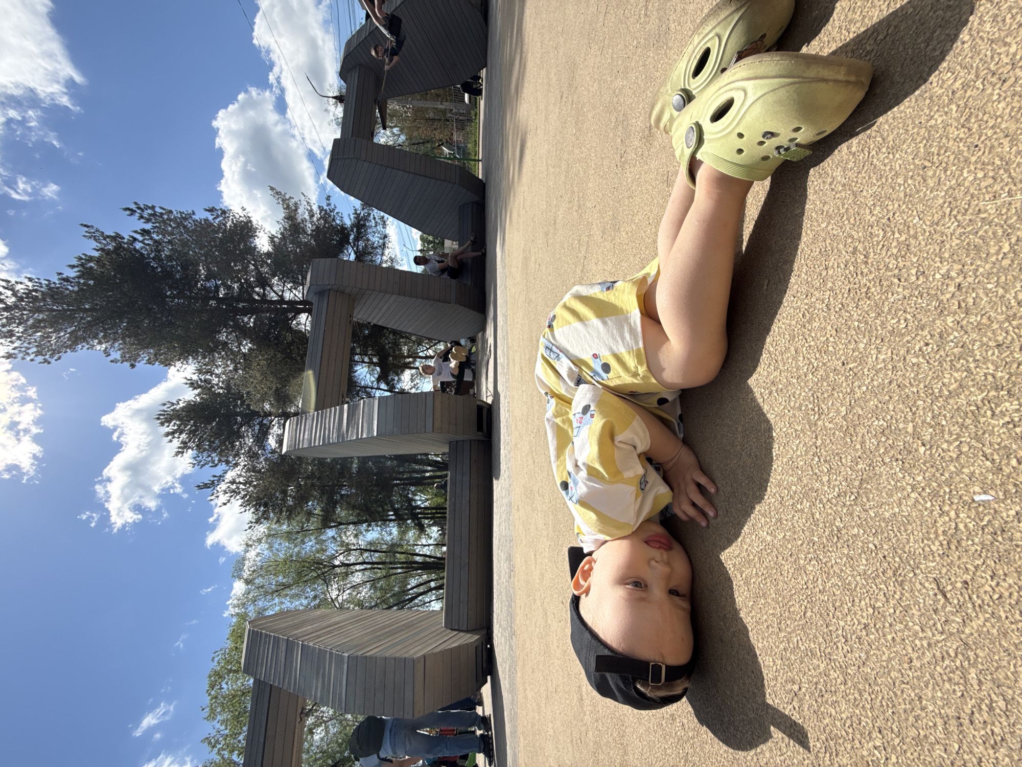 A child is lying on their back on a textured, possibly paved surface in what appears to be a public space. They are wearing light green Crocs shoes and a yellow and white patterned outfit with a black baseball cap. The child is positioned with their head towards the lower-right of the frame.
Behind the child, towards the upper-left of the frame, is a modern, geometric structure made of gray wood. People are visible on and around this structure, suggesting it might be a play area or public installation. The sky is visible in the background, with scattered clouds and trees.
The photo was taken in the city of Rusanovo, Russia.