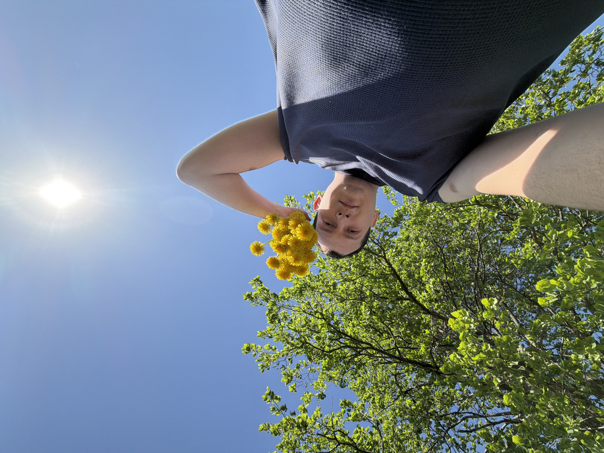 The image shows a man holding a bouquet of yellow dandelions. He is wearing a dark blue t-shirt. The background includes a clear blue sky, the sun, and a tree with green leaves. The location in the city of Кемуль, Russia is not determinable from the image.