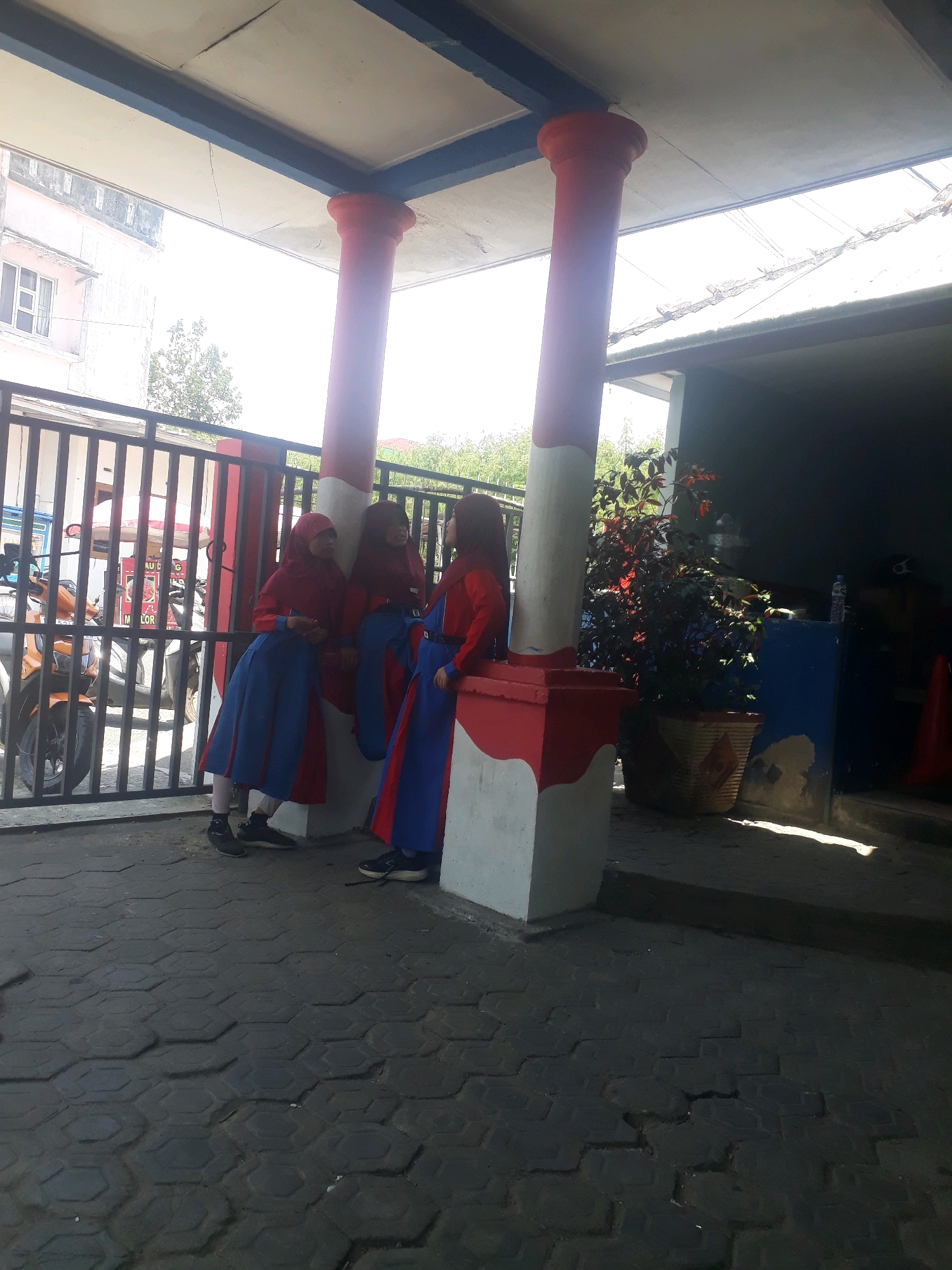 The image depicts three young females in Islamic school uniforms, consisting of red headscarves and blue and red skirts and tops, standing near two large, red and white painted columns. They are positioned on a dark, hexagonal-tiled patio in what appears to be an outdoor area with a metal fence in the background. To the left of the frame, a portion of a building with windows is visible. Behind the fence, a motorcycle is parked. To the right of the columns, a potted plant with dark foliage is situated next to a blue and white structure. The scene is illuminated by bright sunlight, casting shadows on the ground. The image was captured in Pangkalpinang, Indonesia.