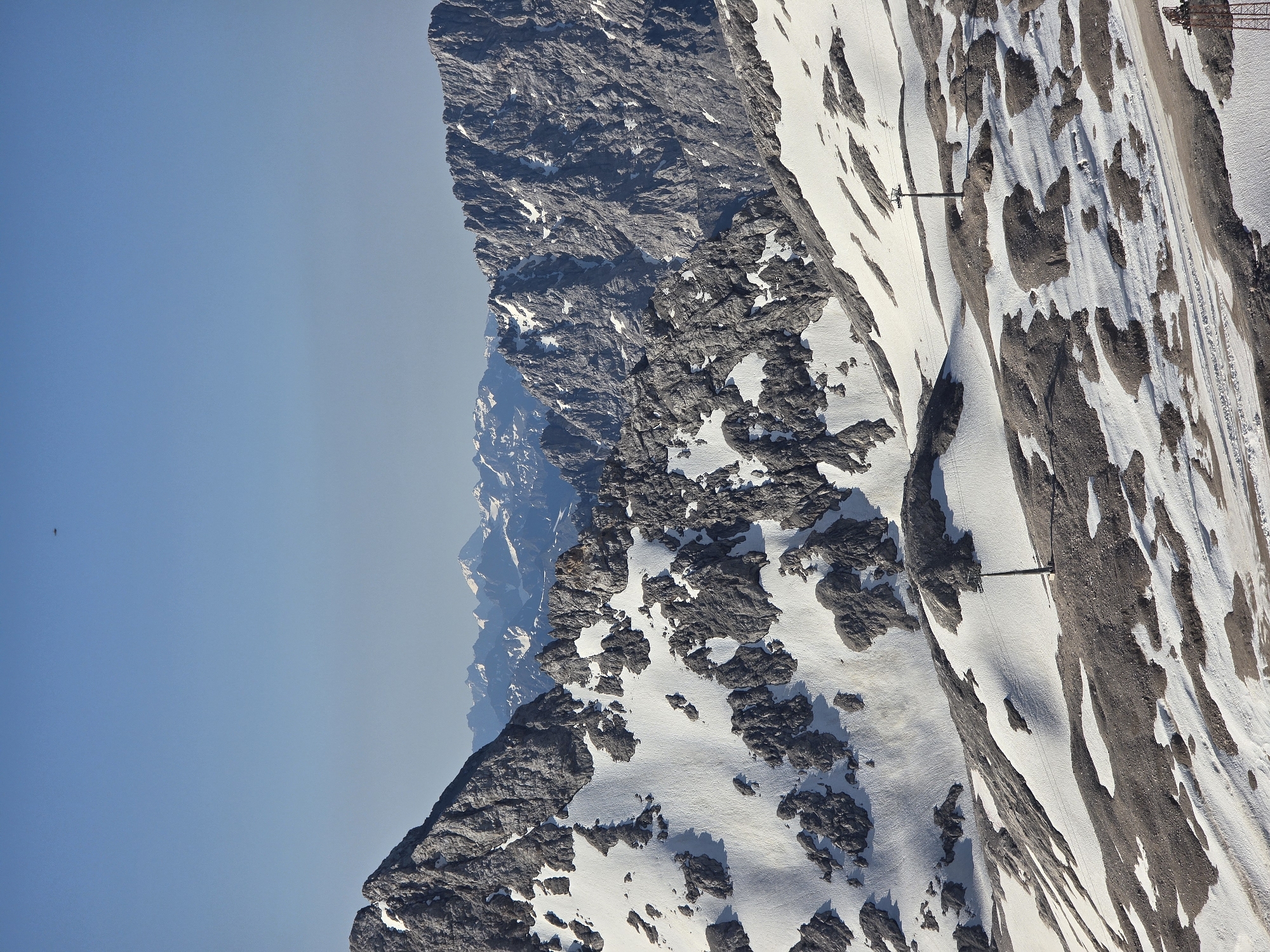 This image captures a panoramic view of a snow-dusted mountainous region, likely part of the Bavarian Alps near Garmisch-Partenkirchen, Germany. The scene is dominated by rugged, rocky peaks partially covered in snow, with a clear blue sky overhead. In the foreground, a ski lift infrastructure is visible, with towers and cables traversing the snowy terrain. The landscape is characterized by steep slopes, scattered rocks, and patches of snow, indicating a high-altitude environment. The distant mountains are also capped with snow, suggesting a vast alpine expanse. A small, dark object, possibly a bird or an aircraft, is visible in the far distance against the blue sky. The overall impression is one of a serene and imposing natural landscape, typical of alpine regions.