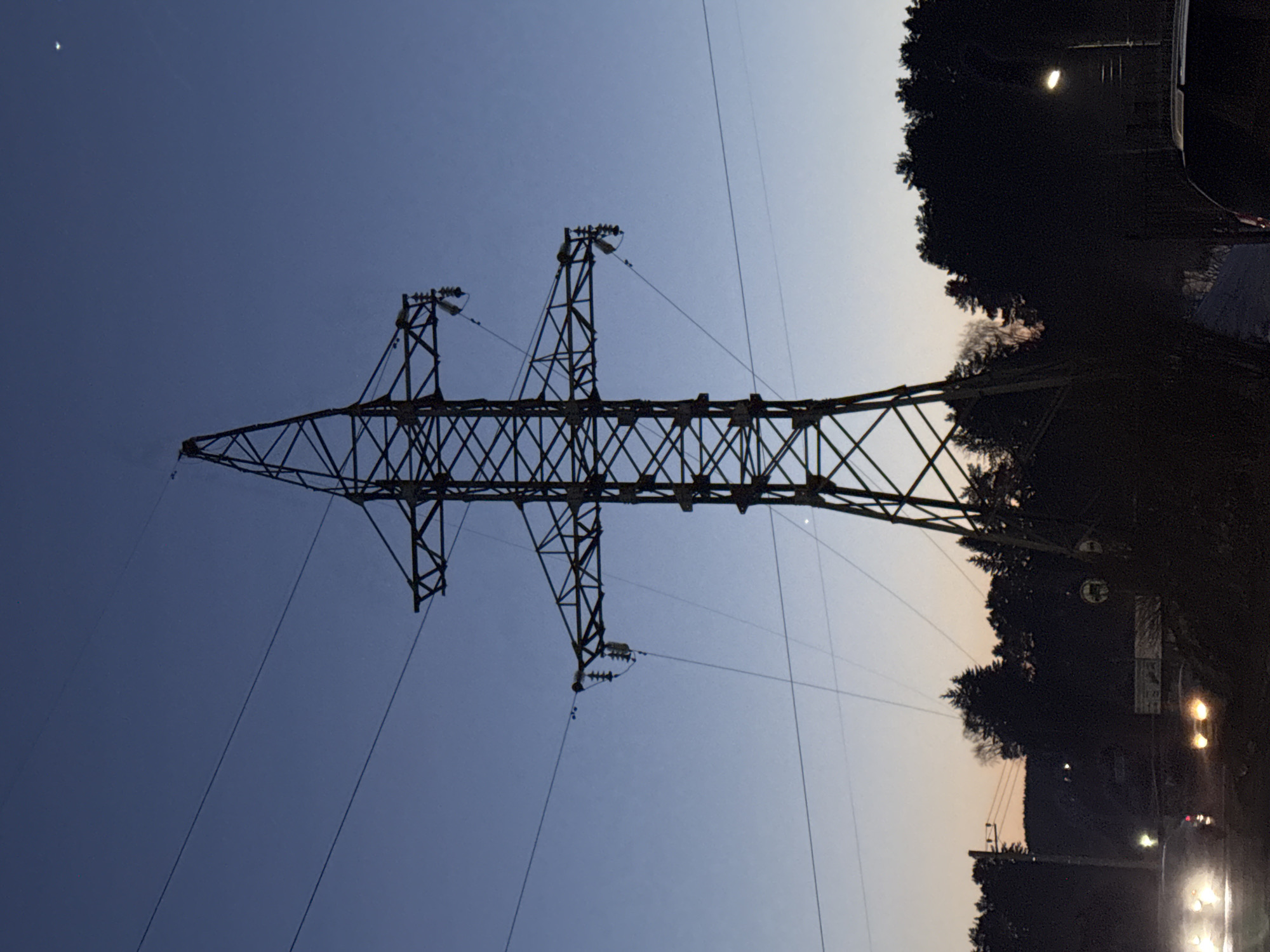 The image shows an electrical transmission tower in Chaikovsky, Russia. The tower is a steel lattice structure, silhouetted against a twilight sky. Thin electrical wires are suspended from the tower. In the background, there's a dark treeline, streetlights, and the blurred lights of passing vehicles.