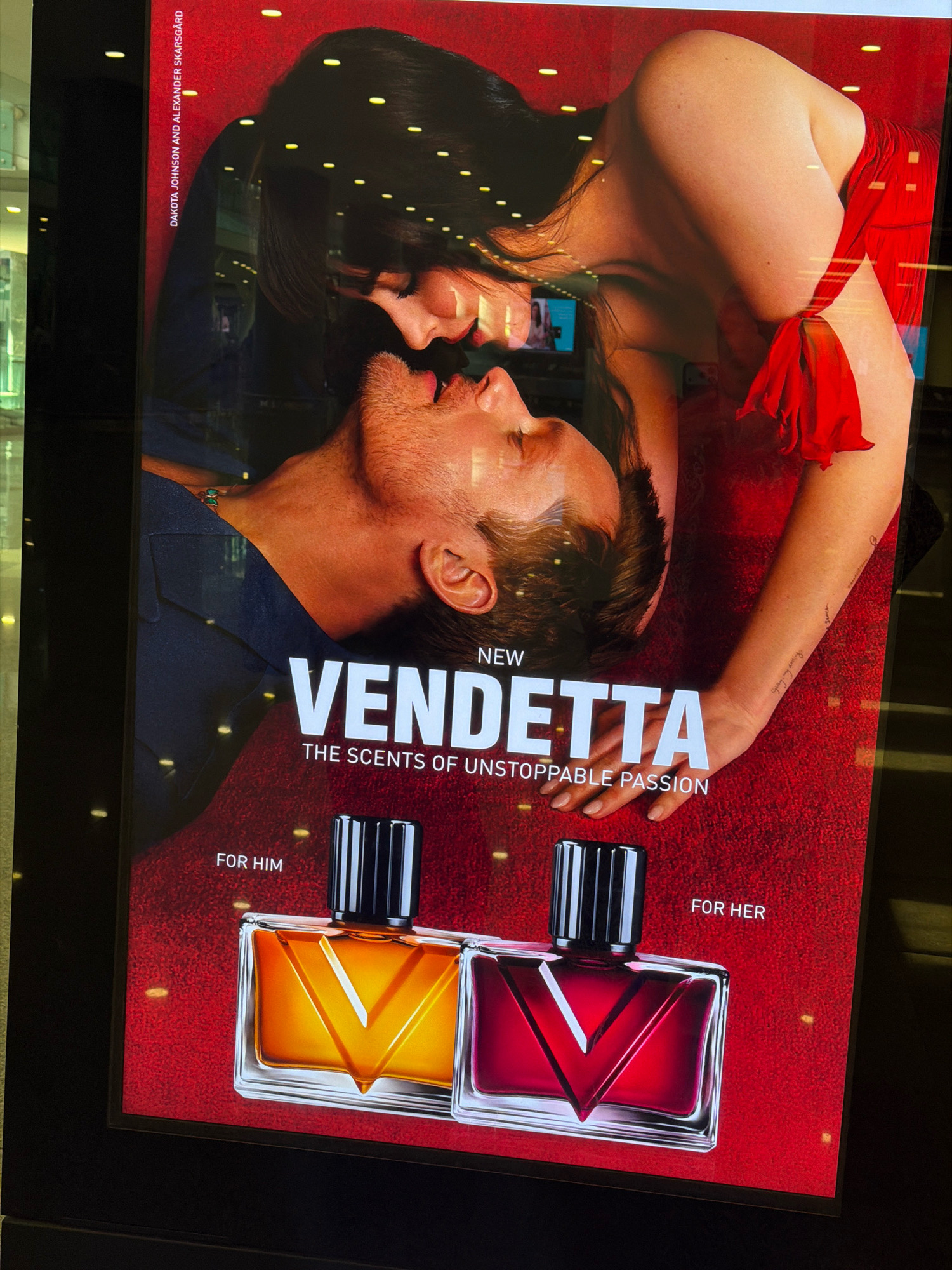 This is a close-up, eye-level, medium shot of a digital advertisement for a perfume called "Vendetta." The ad features a man and woman in an intimate embrace, with the woman's head resting on the man's chest. The background is a textured red surface with scattered yellow lights. The text "NEW VENDETTA THE SCENTS OF UNSTOPPABLE PASSION" is prominently displayed. Below this, two perfume bottles are shown: one amber-colored labeled "FOR HIM" and a red one labeled "FOR HER." The advertisement appears to be in a public indoor space, possibly a shopping mall, with reflections visible on the surface of the display. The image was taken in Gaziemir, Turkey, likely in a commercial setting.