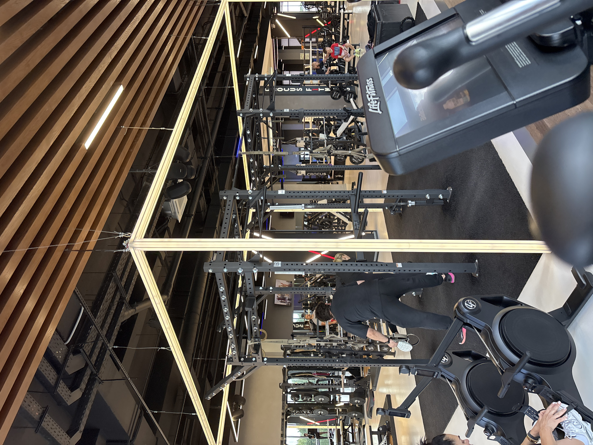 The image depicts the interior of a modern, well-lit gym. The ceiling features a prominent wooden slat design, with recessed lighting and exposed industrial elements like conduits and ventilation systems. In the foreground, a black Life Fitness exercise machine is partially visible, with its screen displaying some content.
The main area of the gym is filled with various weightlifting equipment, including squat racks, weight benches, and dumbbells. A large, gray metal rig with multiple pull-up bars and attachment points stands in the center of the frame.
Several people are present in the gym. In the mid-ground, a person wearing a black t-shirt and black pants is seen from the back, seemingly cleaning or adjusting equipment. To their right, another individual in a red shirt is seated on a bench. Further in the background, more people can be observed utilizing different exercise machines.
The gym floor is covered with black mats in some areas and what appears to be a lighter-colored, polished concrete or linoleum in others. The overall impression is of a clean, professional, and functional fitness facility. The image was taken in Monolit, Russia.