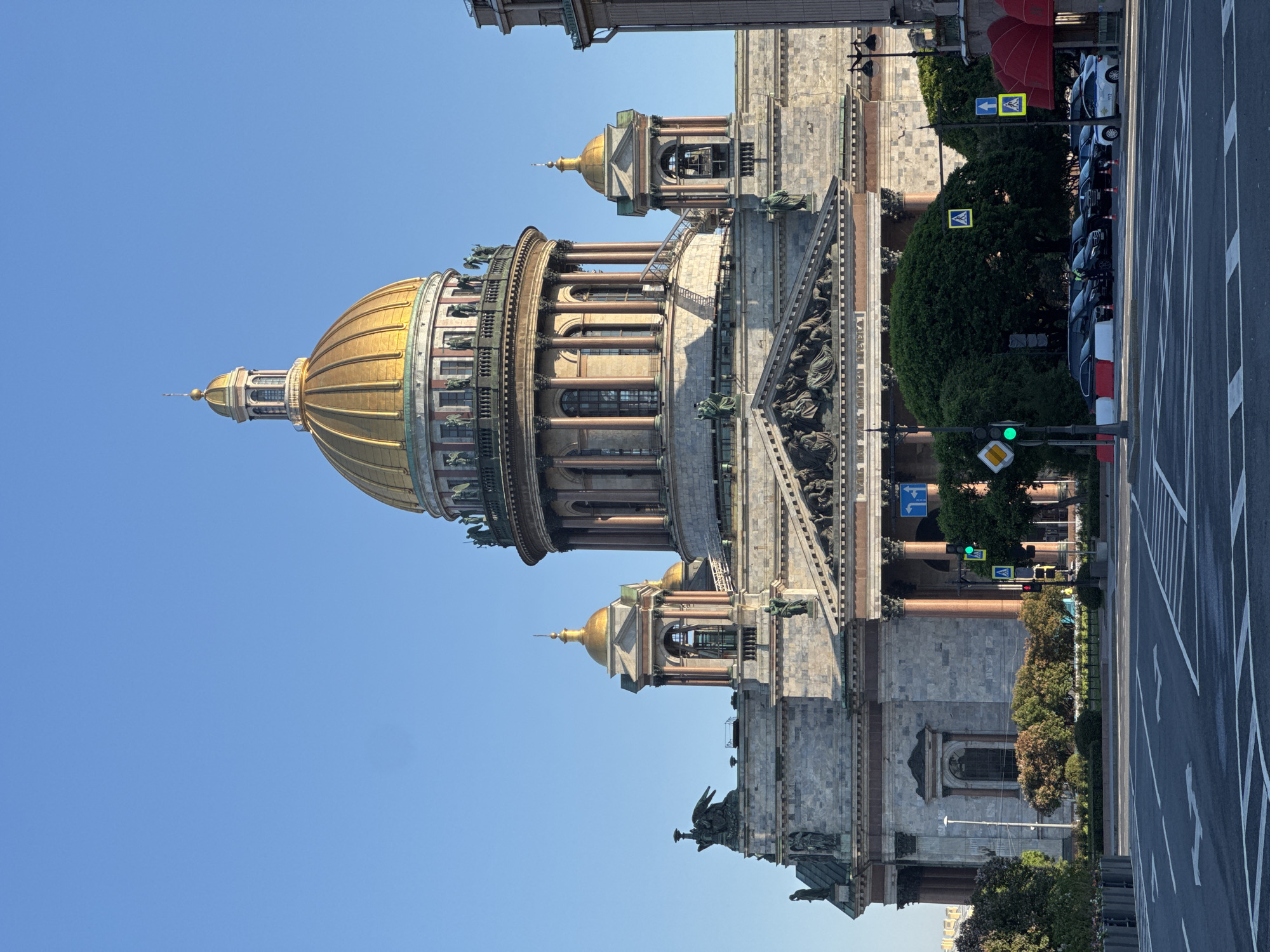 The image displays a grand architectural masterpiece, identified as St. Isaac's Cathedral in Saint Petersburg, Russia, under a clear, bright blue sky. The focal point is the cathedral's massive, ribbed golden dome, which is topped by a smaller cupola and spire. This main dome rests on a drum encircled by numerous columns and arched windows. Flanking the central structure are two smaller golden-domed bell towers or cupolas, also featuring columns.
The cathedral's facade is constructed from large blocks of grey stone, characterized by its neoclassical style with tall columns, intricate sculptural details, and a prominent pediment adorned with relief carvings visible on the right side of the building. Lush green trees and bushes line the base of the cathedral, separating it from the street.
In the foreground, a wide asphalt road with white lane markings stretches across the lower right portion of the image. Several cars are parked along the side of the road. Visible street furniture includes a green traffic light on a pole, indicating an active signal, and various traffic signs: yellow diamond-shaped pedestrian crossing signs with black figures, and a blue rectangular sign with a white right-pointing arrow. The scene is bathed in bright daylight, suggesting a sunny day, likely morning or early afternoon. No people are visible.