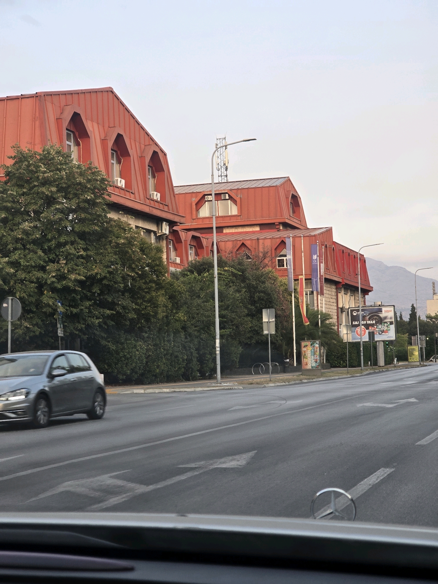 The image shows a street scene in Podgorica, Montenegro. In the foreground, a car's dashboard is visible, with a Mercedes-Benz logo on the hood. The street itself has lane markings and directional arrows painted on the asphalt. To the left, a silver hatchback is in motion, its headlights on. Beyond the street, a large building with a distinctive red, mansard-style roof dominates the scene. The building has multiple dormer windows and appears to be several stories high. Trees and dense foliage line the front of the building, obscuring some of its lower levels. Further down the street, billboards are visible, advertising various products. In the background, mountains can be seen against a pale sky, suggesting a somewhat overcast or hazy day. The overall impression is of a well-trafficked urban environment.