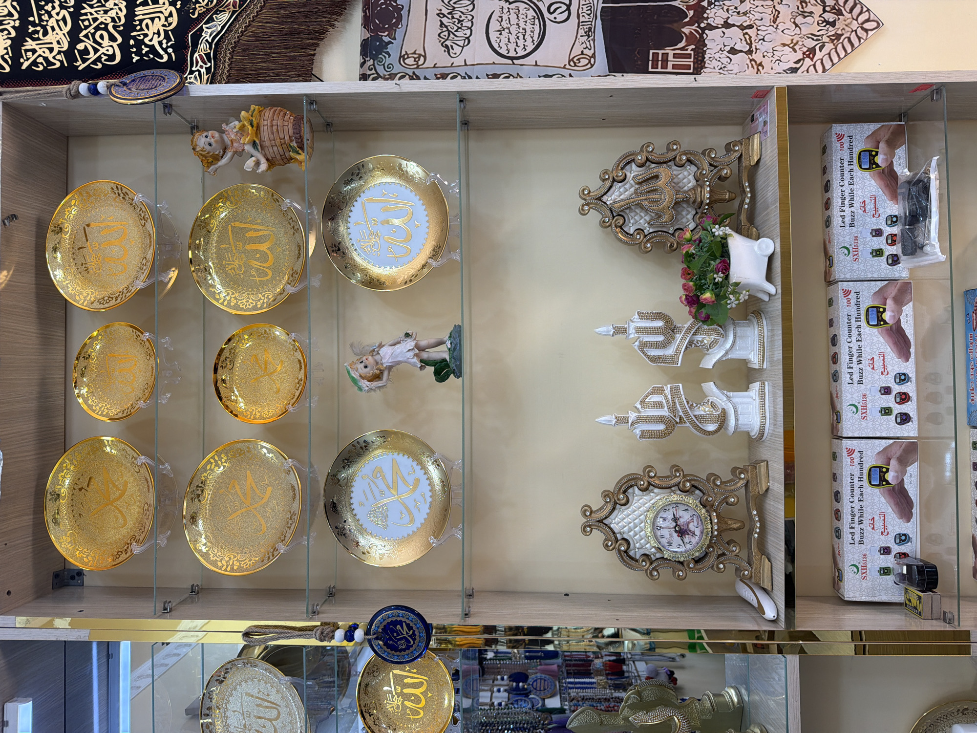 A retail store display in Zangiata, Uzbekistan, features Islamic decorative items on multiple shelves. The shelves contain ornate gold plates with Arabic calligraphy, small figurines of an angel, decorative candles, a clock with an Eiffel Tower design, and a large decorative piece with calligraphy. Several boxes of LED finger counters are stacked on a lower shelf. A tapestry with Arabic script hangs on the right side of the display. The lighting suggests an indoor environment.