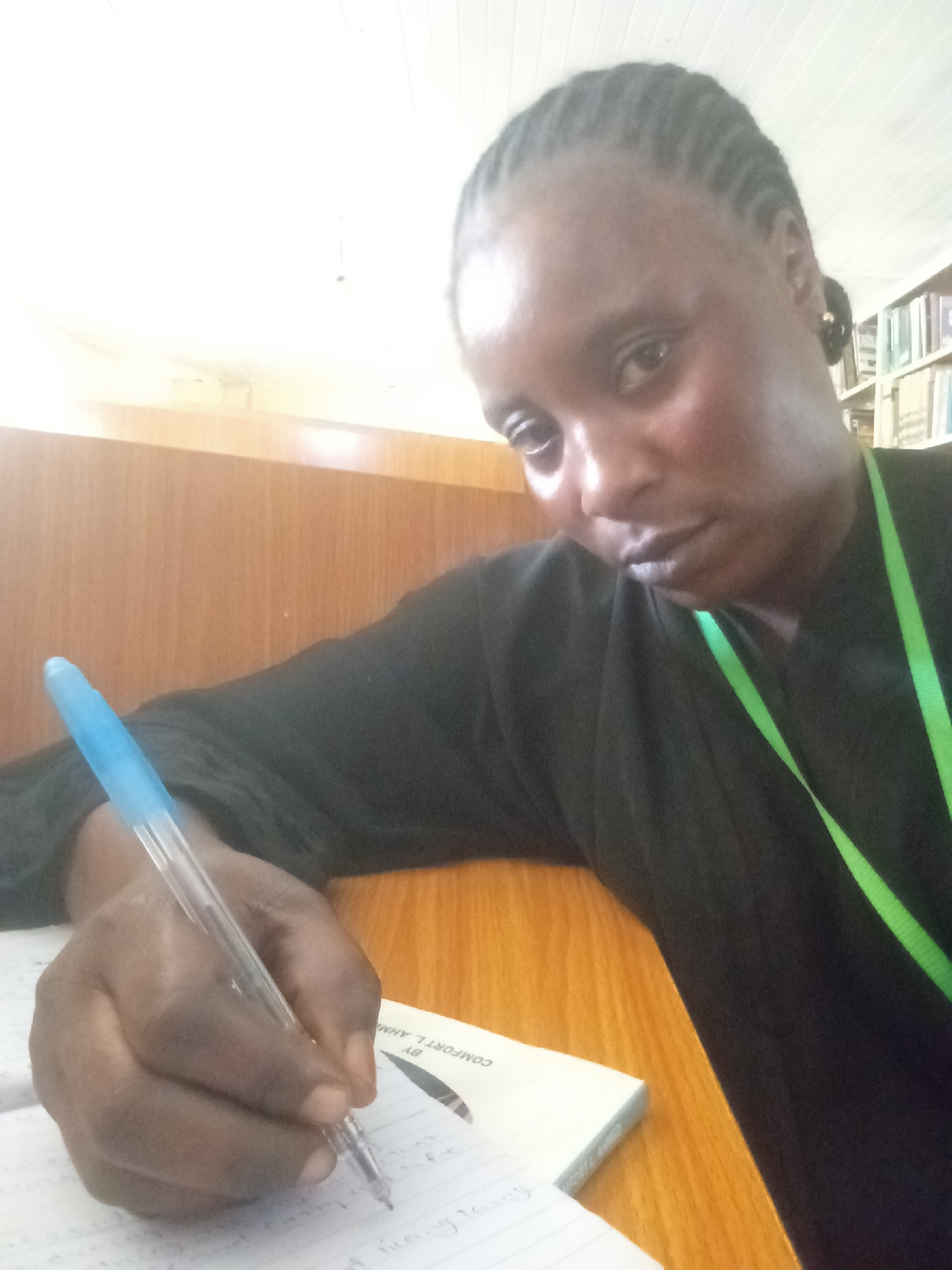 A mid-angle shot depicts a dark-skinned woman writing on a lined notebook with a blue pen. The notebook is partially visible and located on a wooden table. The woman is wearing a black top and a green lanyard. Her hair is styled in braids. A bookshelf is visible in the background. The location is potentially a library in the city of Akwanga, Nigeria.