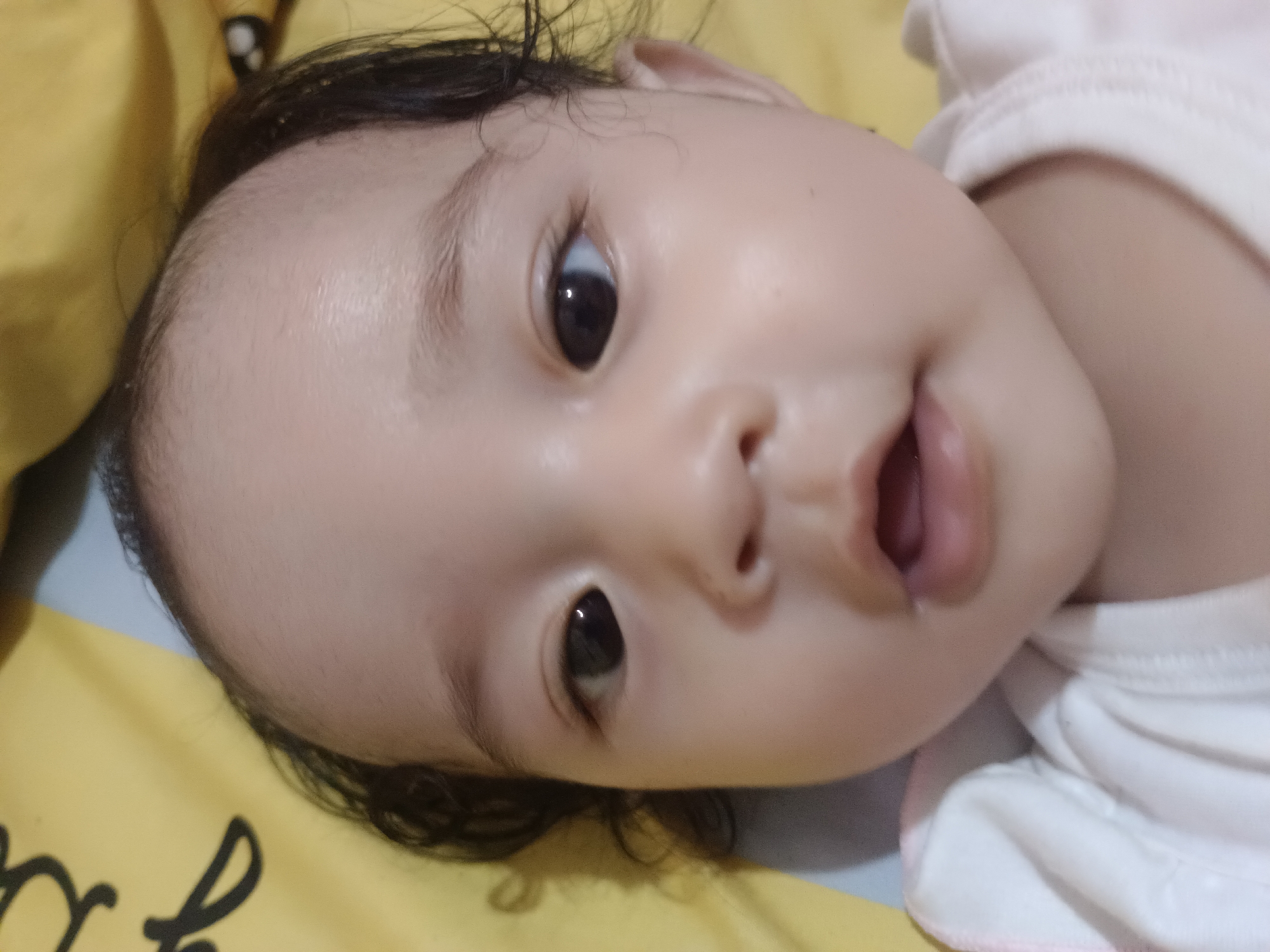 The image is a close-up, horizontally oriented photograph of a baby's face. The baby has dark hair and is lying down on a surface that appears to be a bed with yellow and gray bedding. The baby's eyes are open and looking upwards. The baby is wearing a white garment with pink trim. The focus is on the baby's face, with the forehead and part of the cheek prominently displayed. The lighting is soft and illuminates the baby's skin. The background is largely out of focus, showing hints of yellow and gray fabric. The image does not contain any specific landmarks or contextual clues that would allow for the determination of its location in Kawua, Indonesia.