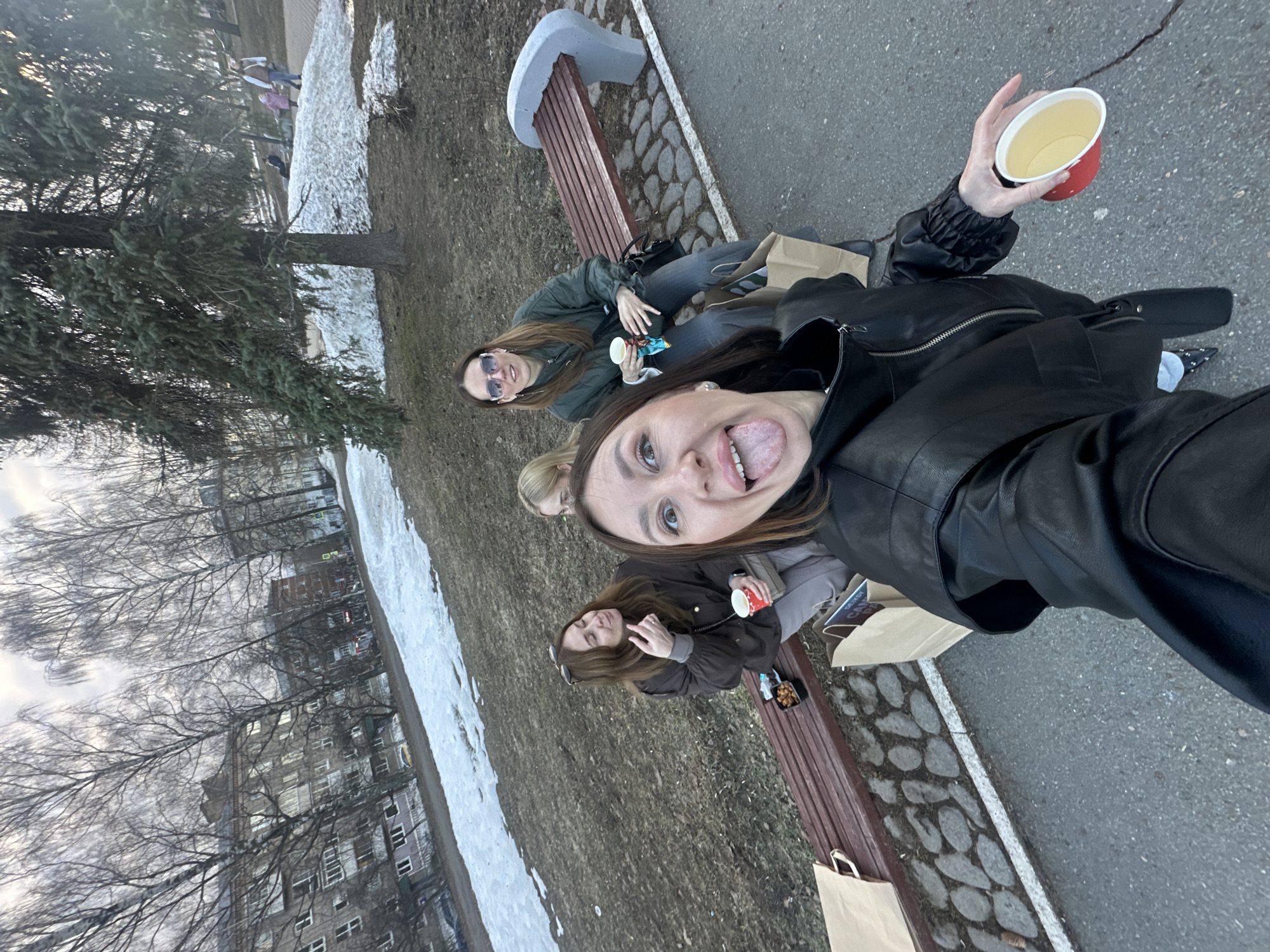 Four women are sitting on a park bench. The scene is likely Izhevsk, Russia. The women are posing for a selfie taken by the woman closest to the camera. The woman taking the selfie is wearing a black leather jacket and sticking out her tongue. The other women are sitting on the bench behind her. There are takeaway bags, and cups on the bench. There is patchy snow on the ground. Buildings and bare trees are in the background.