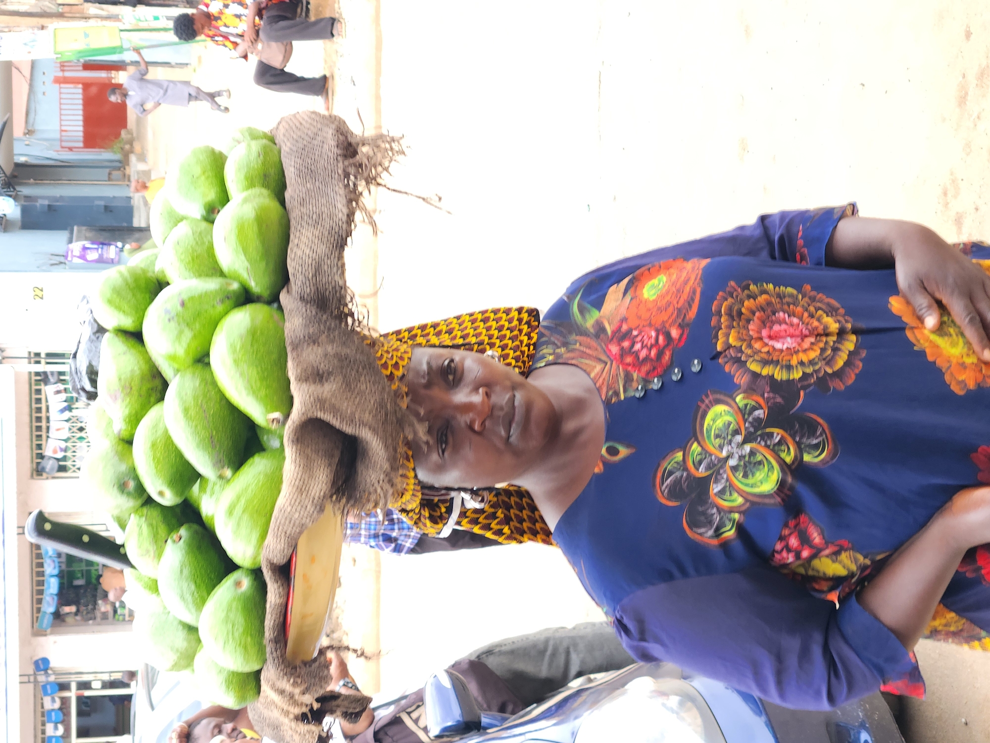 A vibrant and active street scene is depicted, primarily focusing on a woman carrying a large basket of avocados on her head. The woman, positioned prominently on the right side of the frame (when correctly oriented), wears a striking blue dress adorned with large, colorful floral patterns in shades of orange, yellow, and red. A patterned headscarf, yellow and brown, covers her head, providing a base for the woven basket. Her expression is neutral as she looks directly towards the viewer.
The basket on her head is heavily laden with numerous green, ripe avocados, meticulously stacked. Among the avocados, a knife with a black handle is visible, suggesting she might be preparing to cut the fruit or is equipped for sale. A small, yellowish bowl or container is also nestled near the avocados.
In the background, several other individuals are engaged in various activities, contributing to the lively atmosphere of what appears to be a market or public street. One man is seen playing a musical instrument, possibly a drum, while another person in a light-colored outfit appears to be dancing or moving energetically. A partial view of a man's face is also visible at the bottom left.
The setting is outdoors, likely during the daytime, indicated by the bright, even lighting. Buildings with visible details, including the number "22" written on one, form the backdrop. The ground is a light color, possibly dirt or concrete. The overall impression is one of everyday commerce and community interaction in a bustling environment.