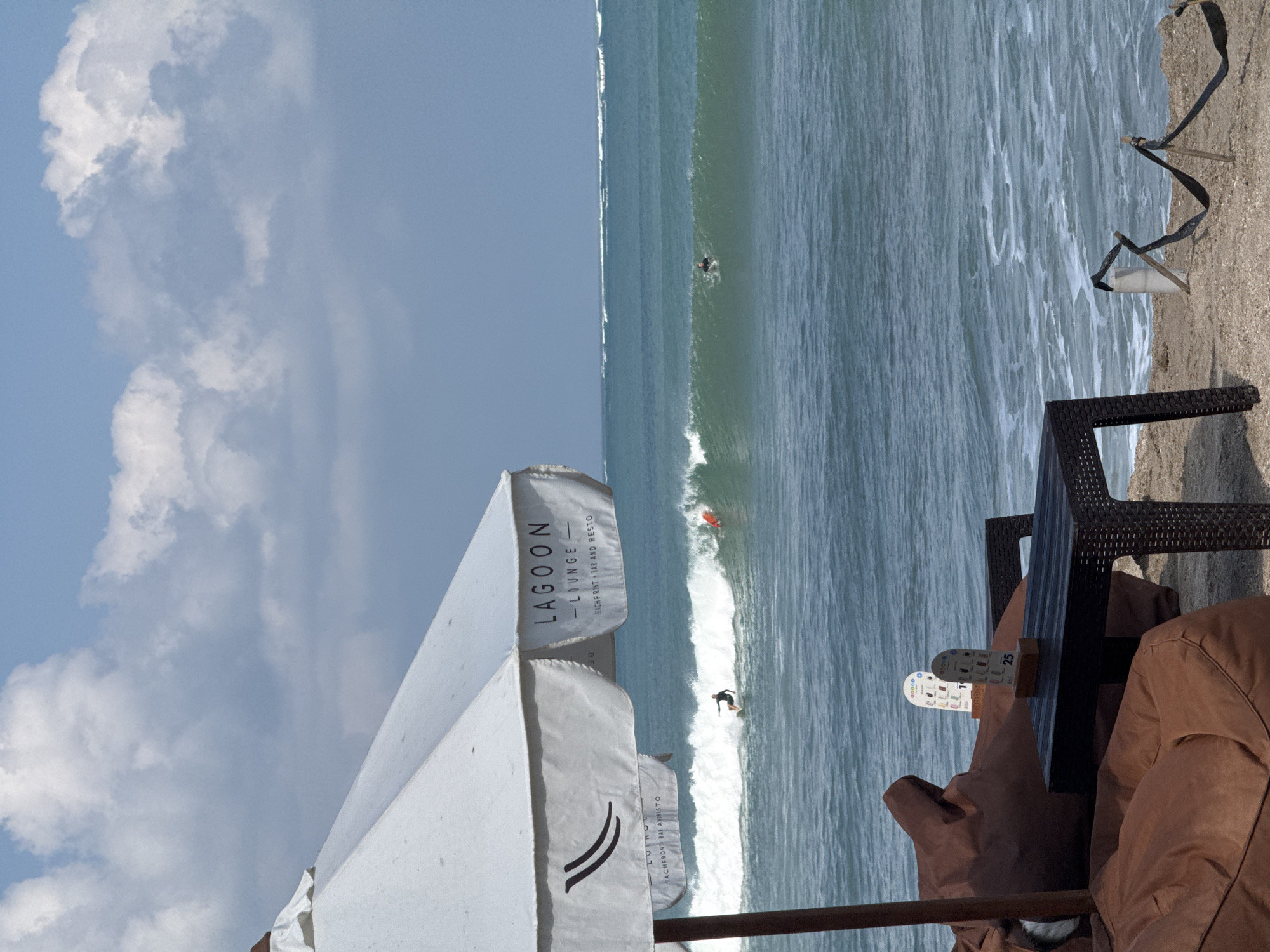 An outdoor seating area faces the ocean in Kuta, Indonesia. A large white umbrella partially shades two large brown beanbag chairs and a dark woven table. Two small menu stands are on the table. In the ocean, waves are breaking, and several surfers are visible, including one riding a wave closer to shore. The sky is partly cloudy. The scene appears to be at a beachfront bar and restaurant, as indicated by the text on the umbrella.