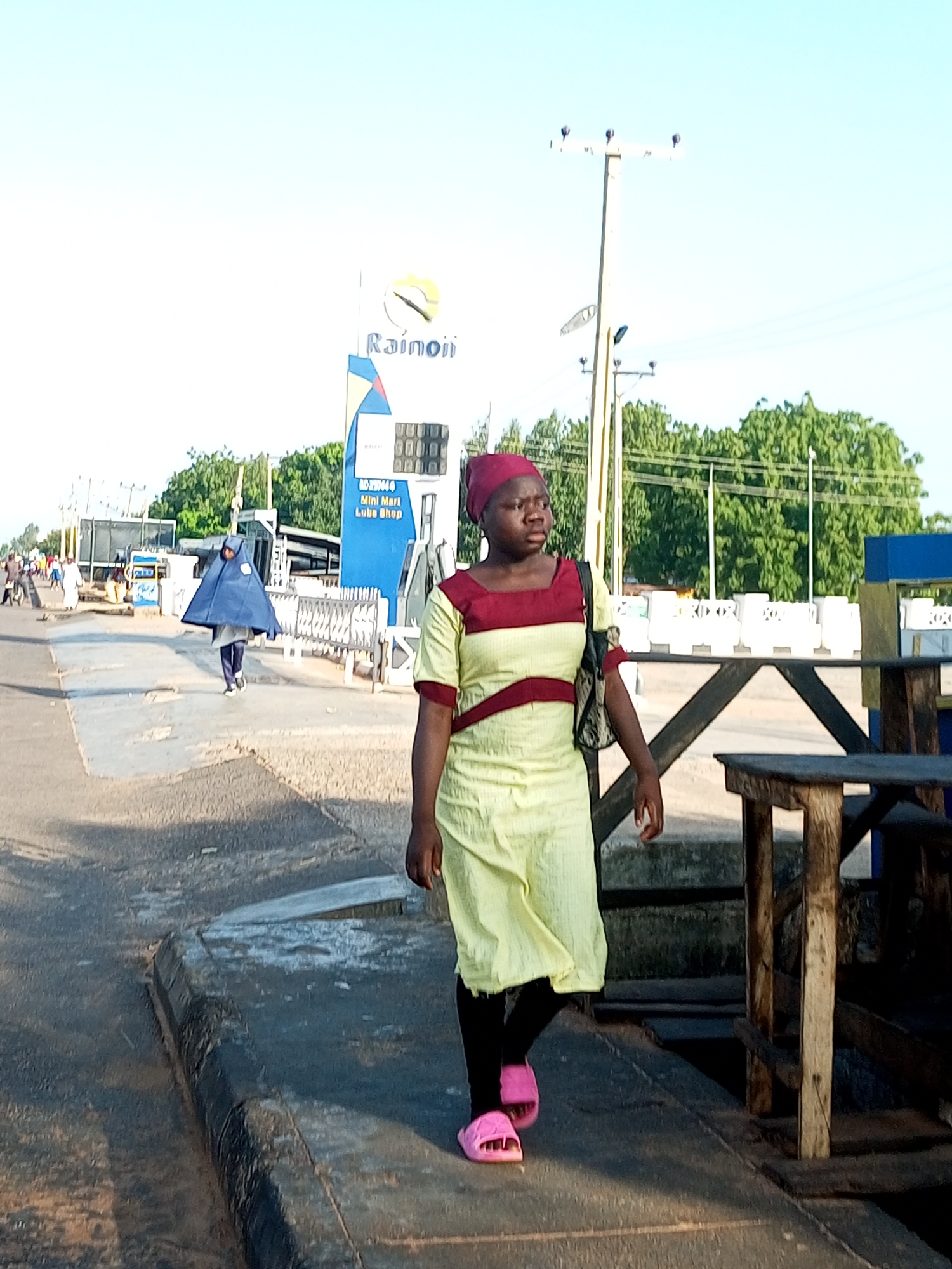 A young Black woman wearing a yellow dress with burgundy trim and a burgundy headscarf walks on a paved area in Garko, Nigeria. She has on black leggings and pink slides. She is carrying a black backpack over her right shoulder. Behind her, a person in a blue hijab walks towards the left. Further in the background, a blue and white sign for "Rainoil" stands tall, with a gas pump and a mini-mart visible. Trees and utility poles line the street, with other pedestrians and vehicles in the distance. The ground is a mix of asphalt and concrete, with a curb in the foreground. The lighting suggests it is daytime with a clear sky.
