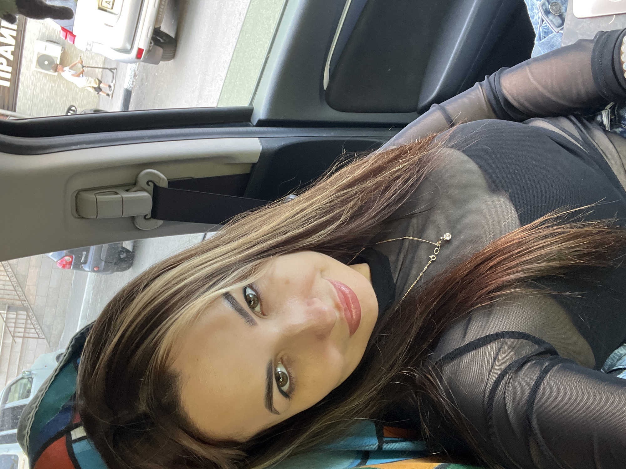A woman is sitting in a car. She is wearing a black, mesh top and has long, brown hair with blonde highlights. A gold necklace is visible. Outside the car, there is a street scene with a pedestrian using a scooter, other vehicles, and buildings. The location is Unknown, Unknown.