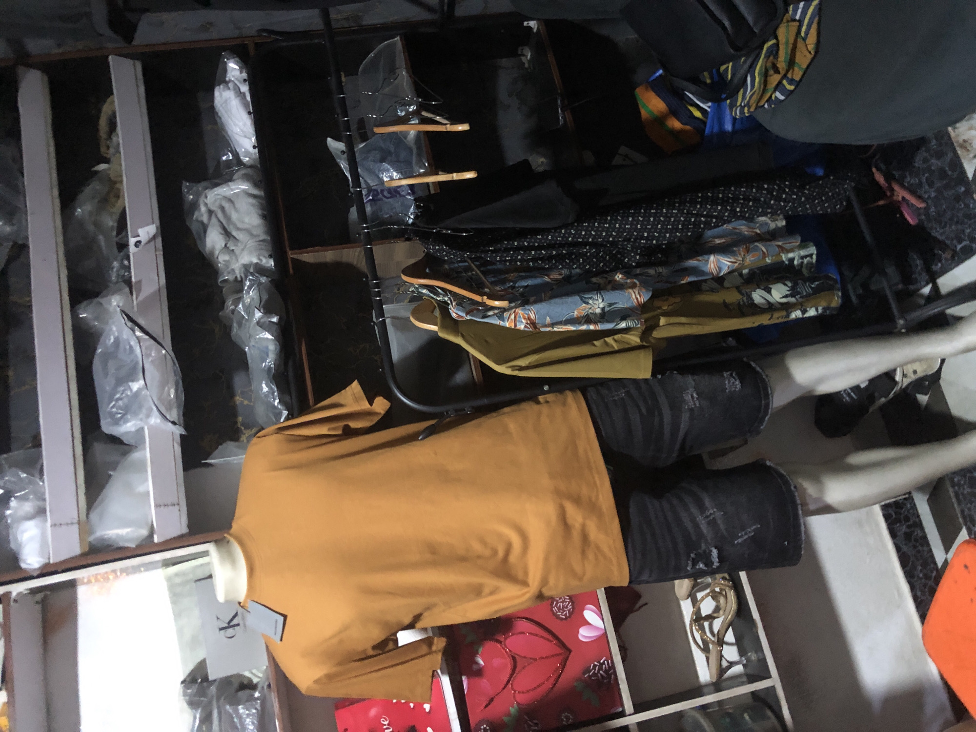 A mannequin wearing an orange t-shirt and dark denim shorts stands in front of a shelving unit in what appears to be a shop in Ado, Nigeria. The shelves are stocked with various items, including clothes in plastic packaging, bags, and a pair of gold strappy heels on a lower shelf. A rack of clothing, including a yellow shirt, a floral print shirt, and a black polka dot dress, hangs to the right of the mannequin. The environment appears to be dimly lit and somewhat cluttered.
