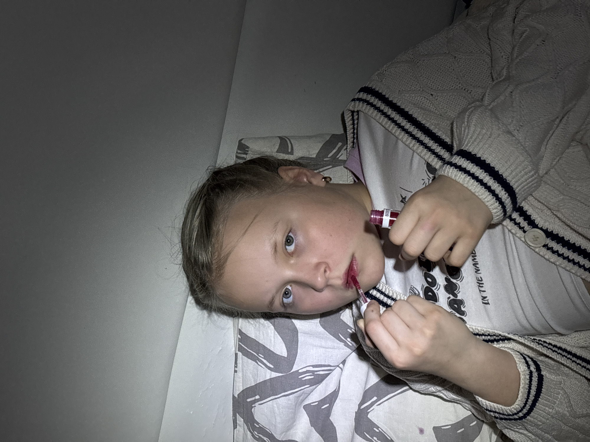 A young, light-skinned female is lying on a bed with a gray-and-white patterned comforter. She is applying red lip gloss to her lips. She is wearing a white t-shirt under a cream-colored sweater with navy blue stripes on the cuffs and collar. The wall to her left is pale gray. There is no evidence to confirm a location in Izhevsk, Russia.