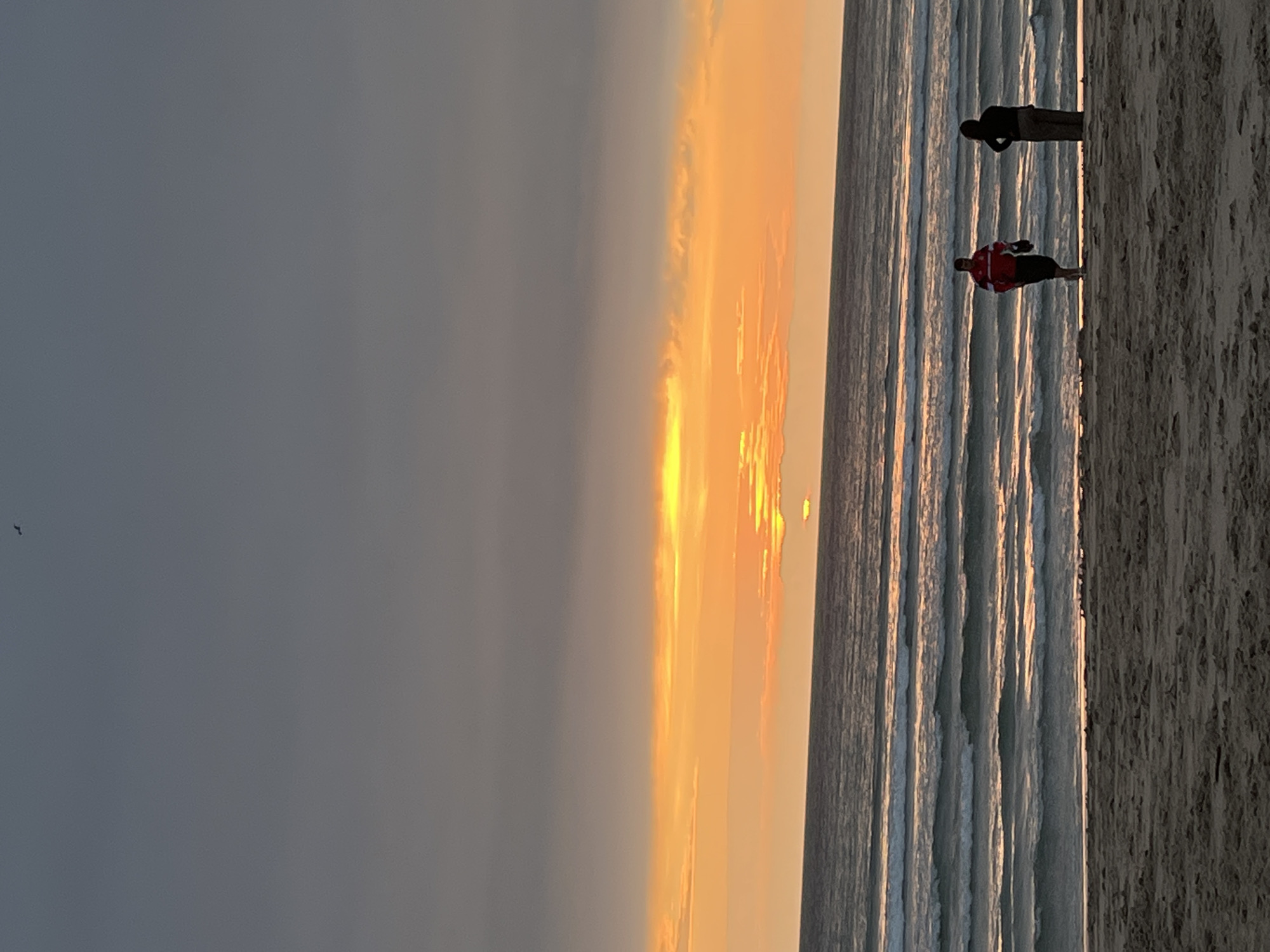 The image shows a beach at sunset in A Coruña, Spain. The sky transitions from dark grey to orange, with the sun low on the horizon. The ocean reflects the sunlight, creating streaks of light. Two people are standing on the beach. One person wears dark clothing and the other wears a red shirt. The beach is sandy.