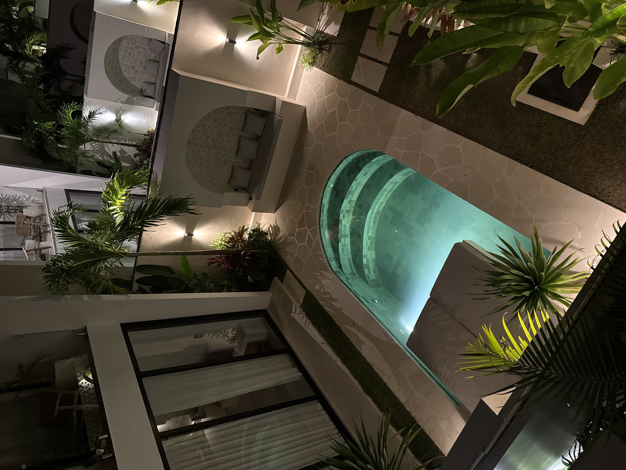 The image captures a nighttime scene in Kuta Selatan, Indonesia, featuring a private pool area. The pool, illuminated by underwater lights, has a curved shape with steps leading into it. A stone-paved area surrounds the pool, with a built-in seating area and cushioned benches along one side. Adjacent to the seating area are tropical plants, adding to the lush environment. Modern architecture is visible, with a building featuring large windows that reflect interior light. The scene is illuminated by outdoor lighting fixtures, creating a warm and inviting ambiance. The photo is taken from an elevated angle, providing an overall view of the space.
