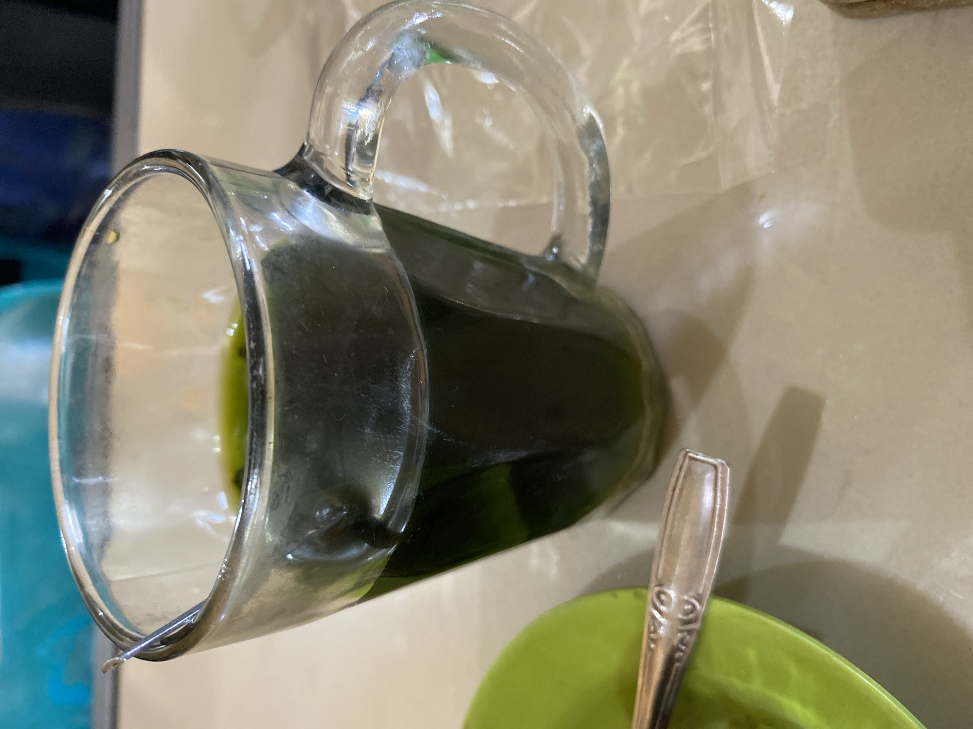 A close-up, top-down, angled shot shows a glass pitcher filled with dark green liquid. The pitcher has a thick, curved handle. The liquid appears to be a beverage, possibly a juice or herbal drink. A shiny metal spoon with an ornate handle is resting on the edge of a bright green bowl, which is partially visible in the lower right portion of the frame. The pitcher is positioned to the left, and its contents create a deep, opaque shadow. The background is a light-colored, textured surface, possibly a tabletop, with a slight sheen that reflects light. A translucent plastic sheet is visible in the upper right corner, creating a hazy overlay. The lighting suggests an indoor setting. No people are visible, and there is no visible text. The scene evokes a sense of refreshment or preparation of a drink. The context of Lhokseumawe, Indonesia, suggests a potentially tropical beverage.