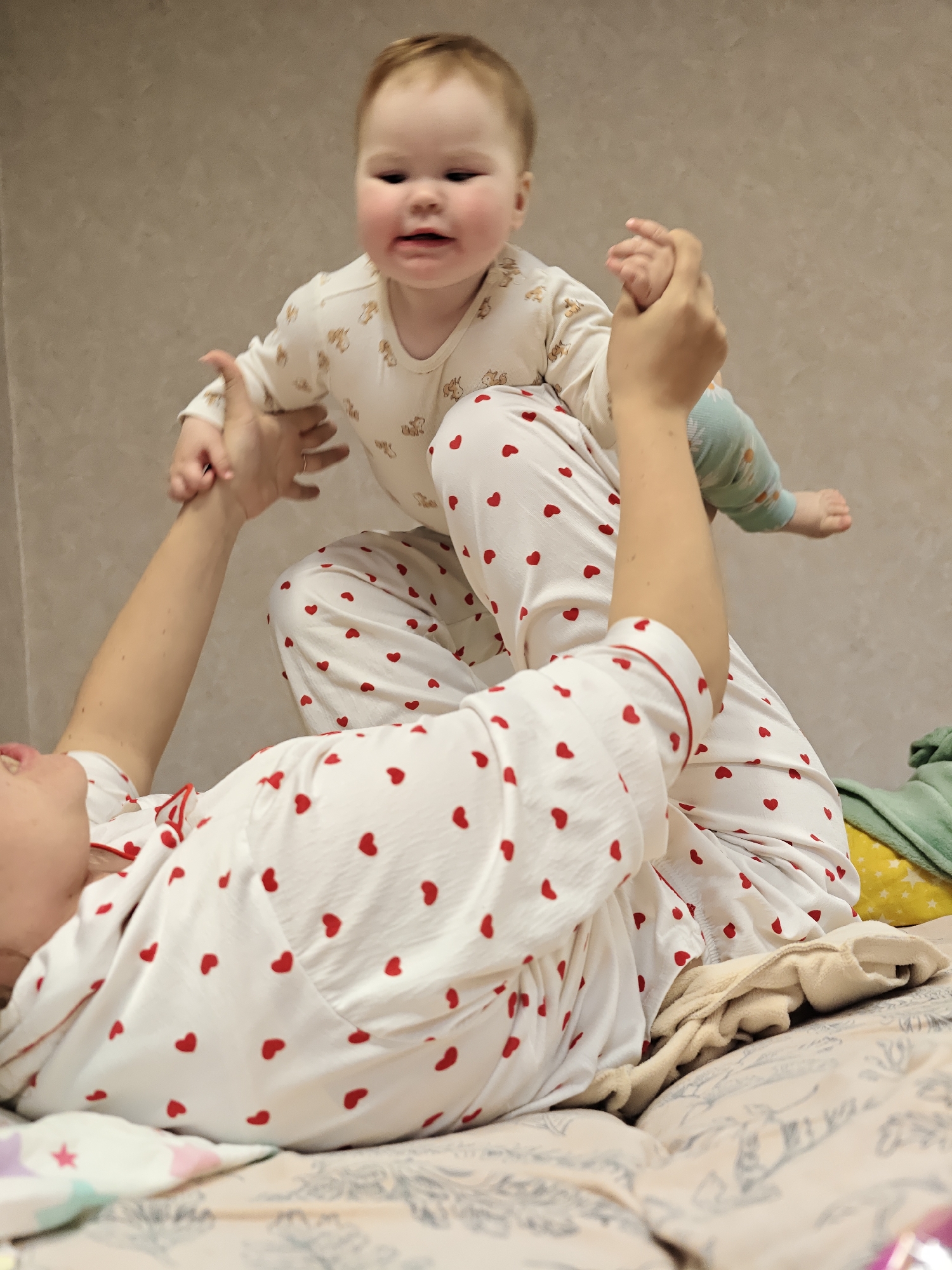 A woman in white pajamas with red hearts lies on her back on a bed. She is holding a baby in the air above her. The baby is wearing a white onesie with a bear pattern and green patterned socks. The background is a beige wall. The image was taken in Chaikovsky, Russia.