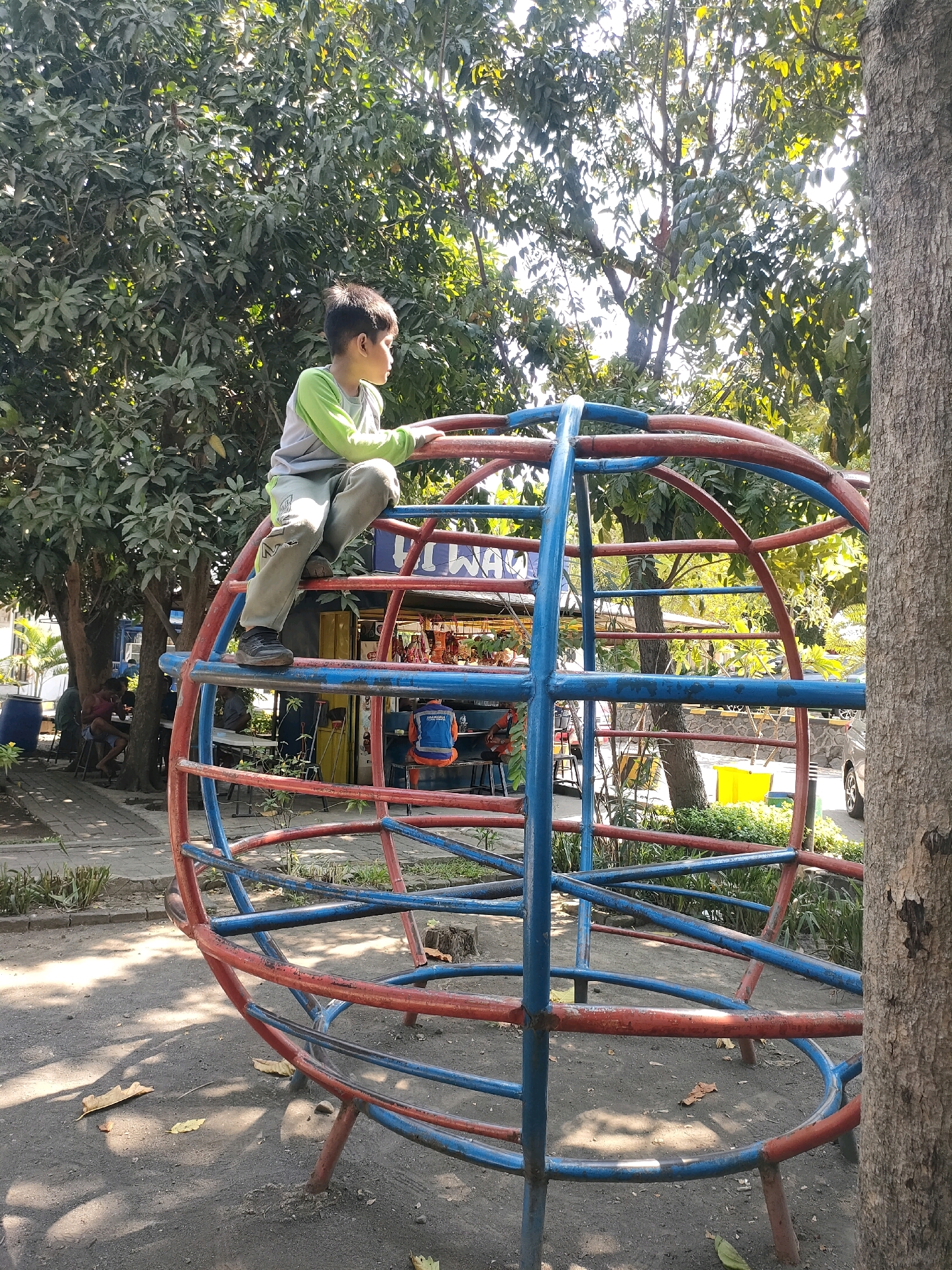 In Sukodono, Indonesia, a young boy with dark hair is perched on a large, spherical jungle gym structure made of red and blue metal bars. He is wearing a light green long-sleeved shirt and light-colored pants with rips at the knees. The boy is looking to his right, off-camera. The jungle gym is situated in an outdoor environment with trees and foliage in the background. The ground beneath the jungle gym is a mixture of dirt and sand, with fallen leaves scattered about. Sunlight filters through the trees, casting dappled shadows on the ground. In the background, other people are visible sitting at tables, possibly in a park or food stall area. A large tree trunk is prominent on the right side of the frame.
