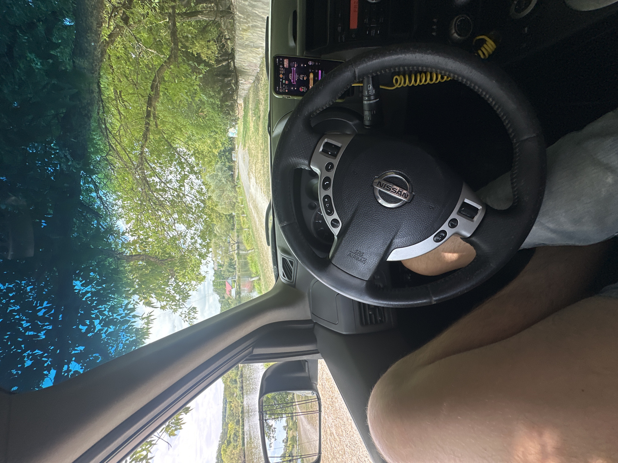 The image depicts the interior of a Nissan vehicle, viewed from the driver's perspective. The driver's leg is visible in the foreground, and the steering wheel, dashboard, and a smartphone mounted on the dashboard are prominent. Outside the vehicle, a dirt road leads towards a body of water, likely a river or lake, bordered by trees and vegetation. The scene appears to be a rural or natural setting. The metadata associated with the image indicates it was taken in Makiv, Ukraine.