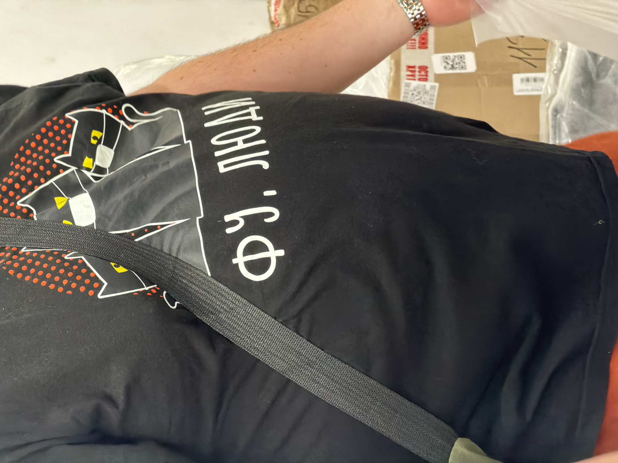 A mid-shot captures the torso and right arm of an individual, located in Saint Petersburg, Russia, against a backdrop of stacked cardboard boxes. The person wears a black short-sleeved t-shirt adorned with a graphic featuring three black cats outlined in white, each possessing yellow eyes. Two of the cats are depicted wearing white face masks. Above the cats, a pattern of red polka dots is visible on the t-shirt. Below the cat graphic, white Cyrillic text reads "ФУ, ЛЮДИ," which translates to "Ugh, people." A black, coarsely woven strap, consistent with a bag strap or seatbelt, traverses diagonally across the person's chest from the upper left to the lower right. The visible portion of the right forearm exhibits light hair, and a metallic, silver-toned link watch is worn on the right wrist. The individual's right hand holds a translucent white plastic bag. The background consists of numerous brown cardboard boxes, many displaying shipping labels, barcodes, QR codes, and fragments of Cyrillic text, including "ОСТО" and "ХРУП" (Fragile), indicating a Russian context. A handwritten number "119" is discernible on one of the boxes. The scene depicts an indoor environment, possibly a storage or shipping area.