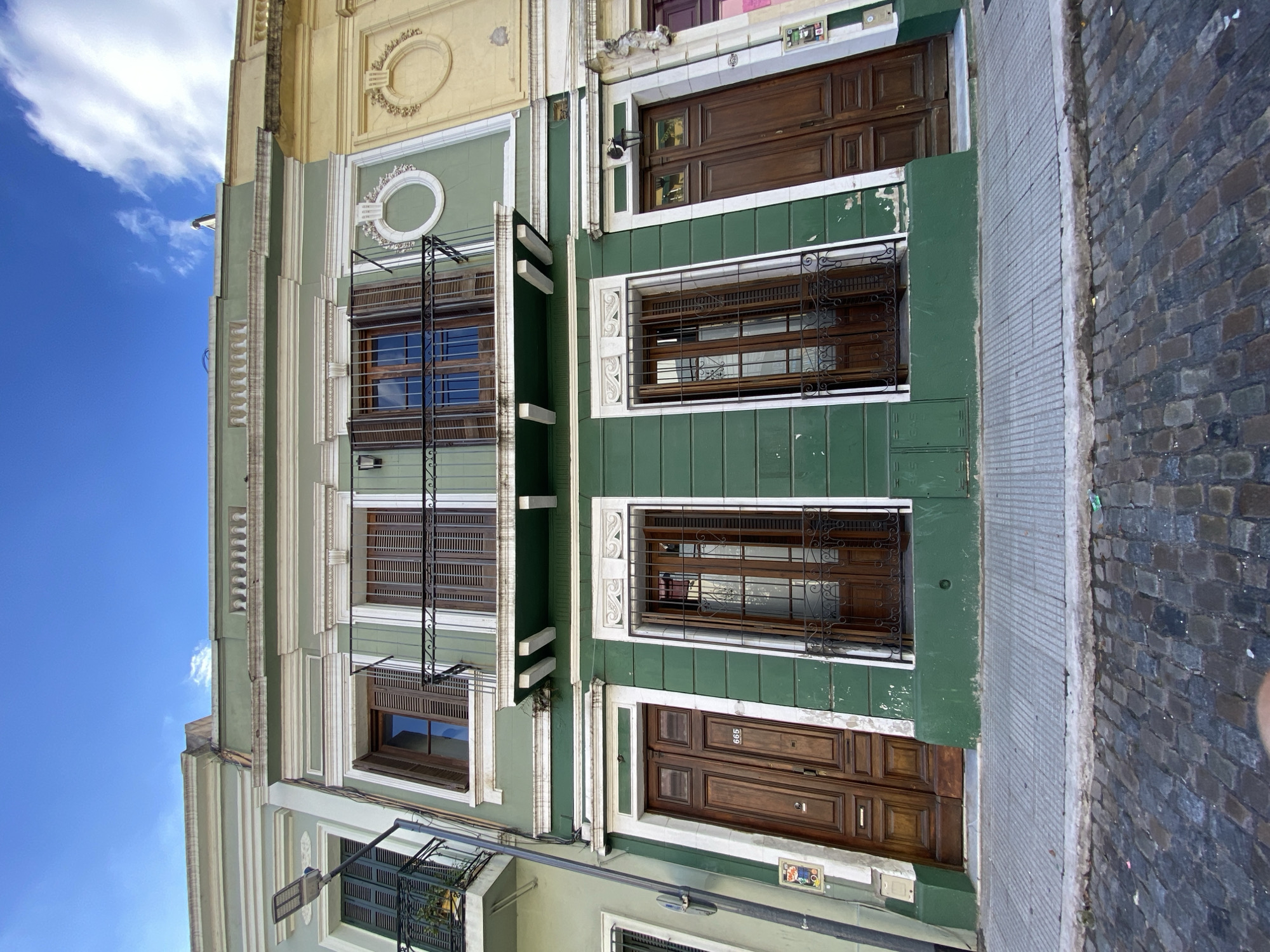 The image shows a building facade in Buenos Aires, Argentina. The building has a light green and white color scheme with dark brown wooden doors and windows. There are wrought iron window bars on some windows. The buildings adjacent to it are painted yellow on the left and green on the right. The street and sidewalk are made of cobblestone. The sky is blue with white clouds.