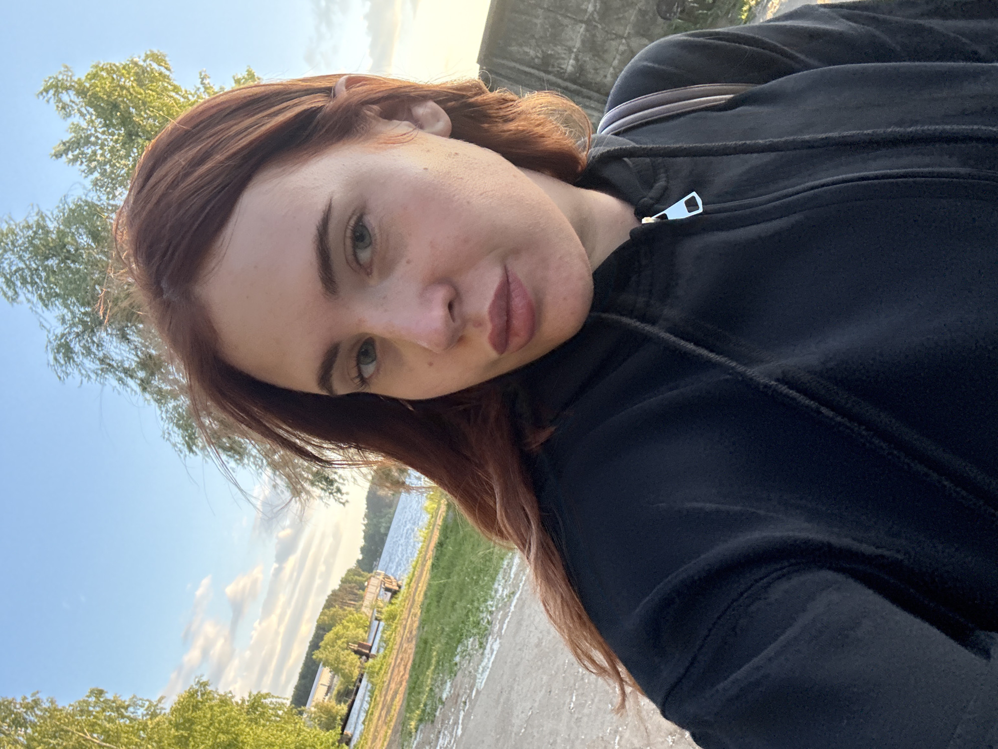 A young woman with auburn hair takes a selfie outdoors, wearing a black zip-up hoodie. She stands with a body of water and lush greenery behind her, with trees and a cloudy sky visible. A concrete structure is to her right, and a path stretches out in front of her. The scene appears to be near the Kama River, within the city of Chaikovsky, Russia, potentially at a waterfront park or recreational area. The lighting suggests it is late afternoon or early evening.
