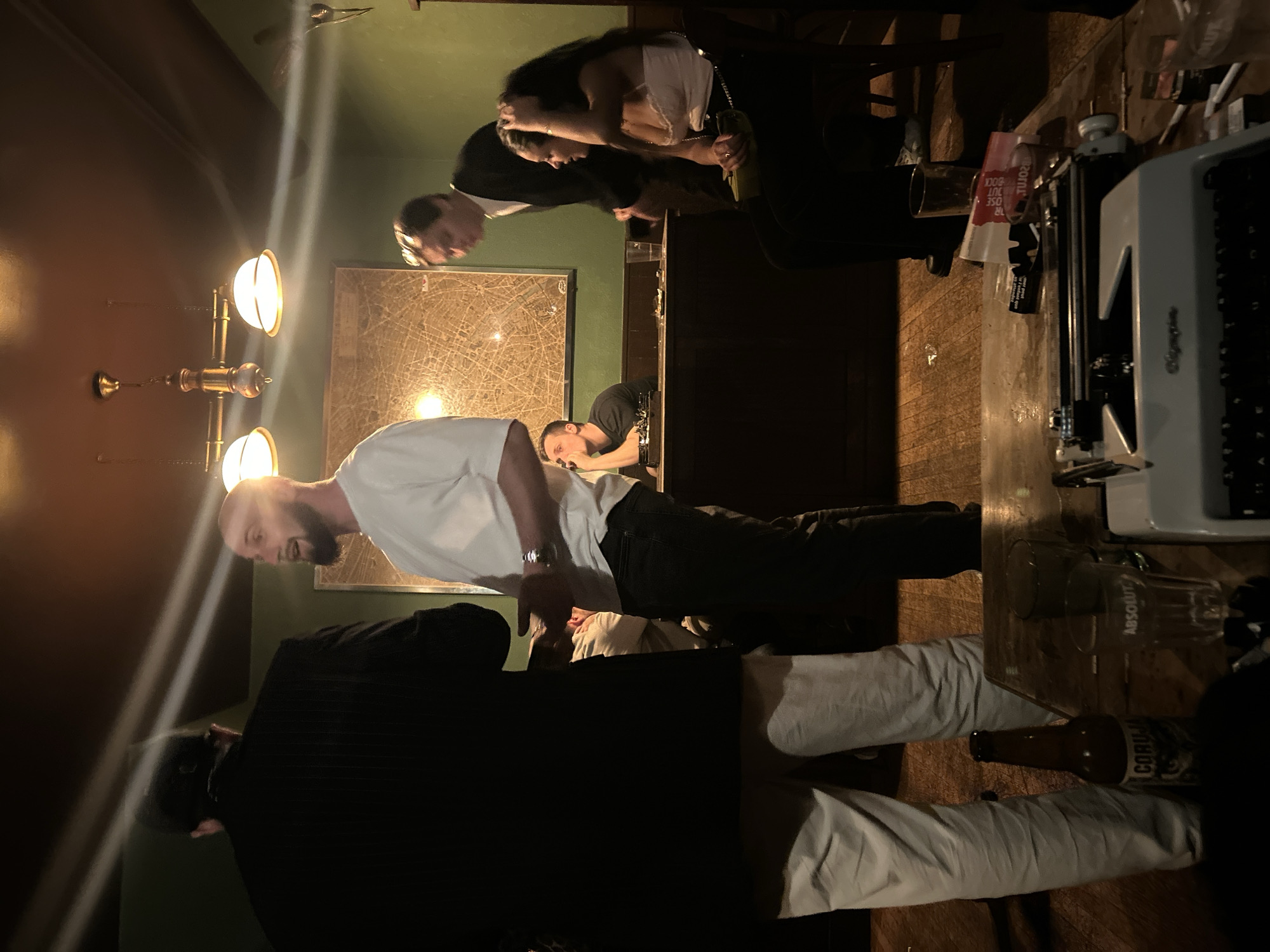 **Scene:** Interior of a dimly lit bar or restaurant.
**People:** Several individuals are present. One man with a shaved head and beard is wearing a white t-shirt and dark pants. Another man is wearing a black blazer and light-colored pants. Two other people are partially visible in the background behind the bar, engaged in conversation. Another person is leaning on the bar further back.
**Objects:** An antique typewriter rests on a wooden surface. There's a glass labeled "ABSOLUT" and a bottle of "CORONA" beer. A large framed map hangs on the green wall. An ornate chandelier with multiple lights illuminates the scene.
**Actions:** The people are standing and appear to be engaged in conversation.
**Interactions:** Some people are directly interacting near the bar.
**Environment:** The interior has a warm color palette with dark wood and green accents. The lighting is low and focused, creating a cozy atmosphere. The setting evokes a traditional or vintage-inspired establishment. There is no evidence to suggest the location is in Paris, France.