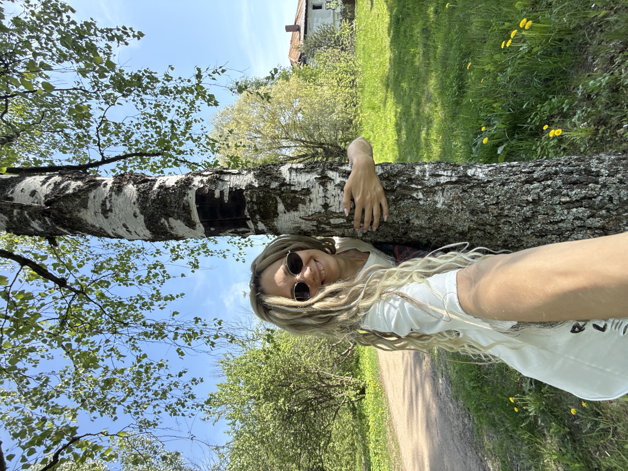A woman is posing for a selfie next to a tree. She is wearing sunglasses and a white t-shirt. The background consists of trees, grass, and a house in the distance. The photo was taken outside during the day in the city of Unknown, Unknown.
