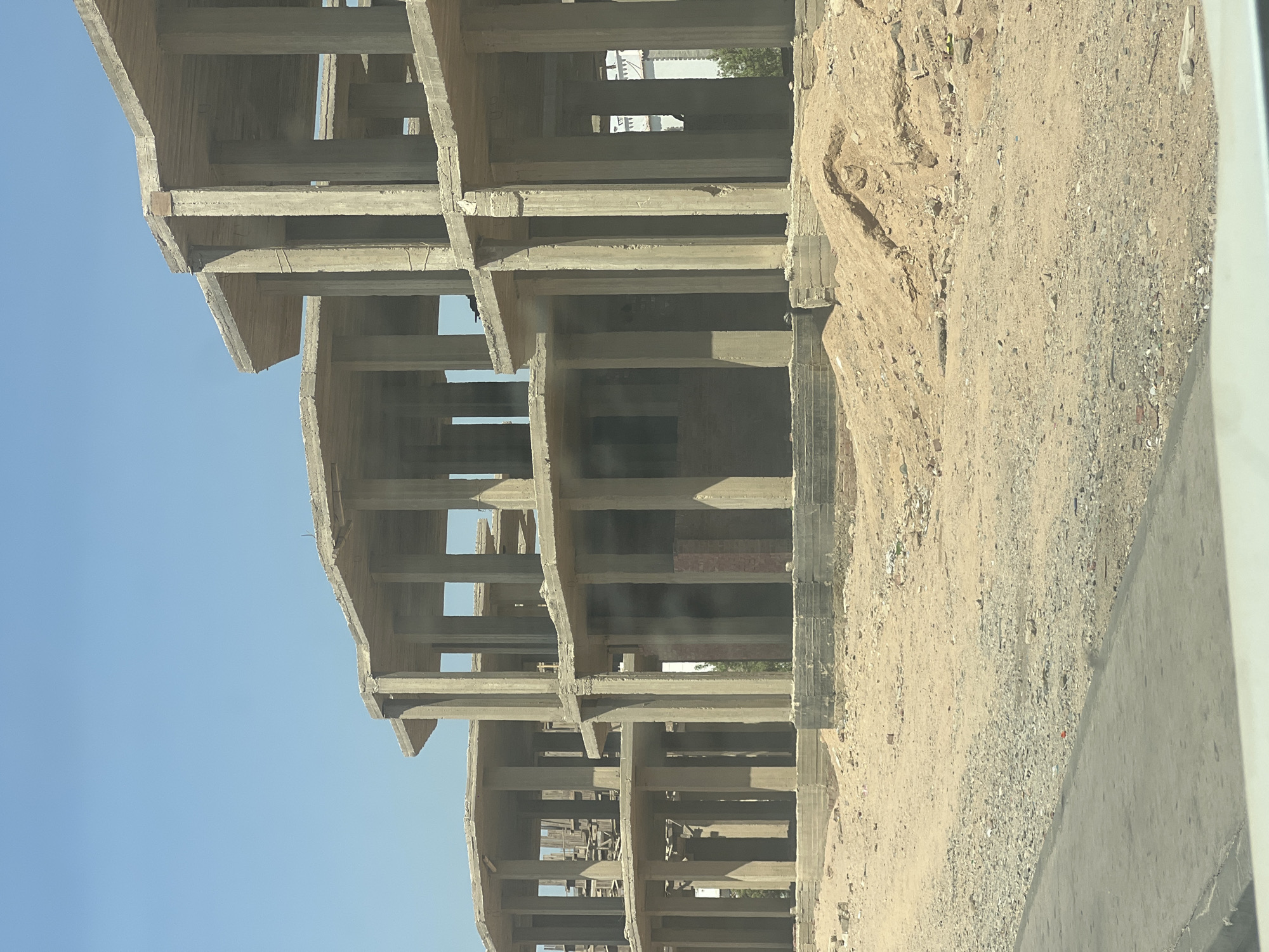 The image shows a construction site in Sharm El Sheikh, Egypt, featuring the unfinished concrete skeletons of several multi-story buildings. The structures are characterized by exposed concrete columns and slabs, with open spaces where walls and windows would typically be. The surrounding environment is arid and dusty, with piles of sand and gravel on the ground. The sky is clear and blue, indicating a sunny day. There are no people visible in the frame, and the focus is solely on the architectural structures and the desolate landscape. The scene suggests an ongoing construction project that is either in its early stages or has been halted.
