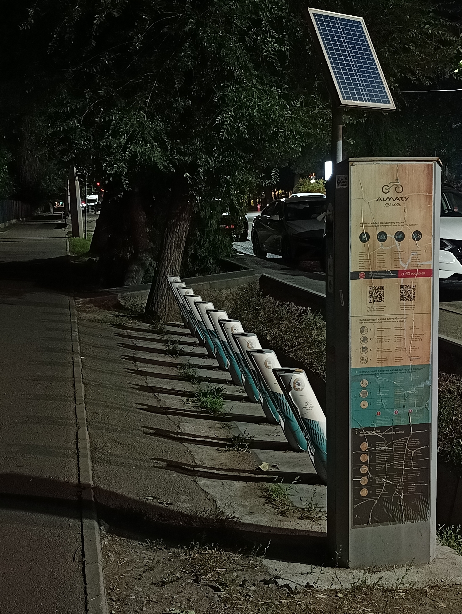 This is a nighttime, medium-shot photograph of a public bike-sharing station in Almaty, Kazakhstan. The station features a row of white bike racks with teal accents. A prominent solar panel is mounted on a pole above the information kiosk, which displays instructions and a map in what appears to be Kazakh and Russian, with some English translations. The kiosk has QR codes for access and contact information. A sidewalk and street are visible to the left, with a large tree casting shadows. Several cars are parked in the background. The scene is dimly lit by streetlights and the solar panel, creating a somewhat stark and urban atmosphere.
