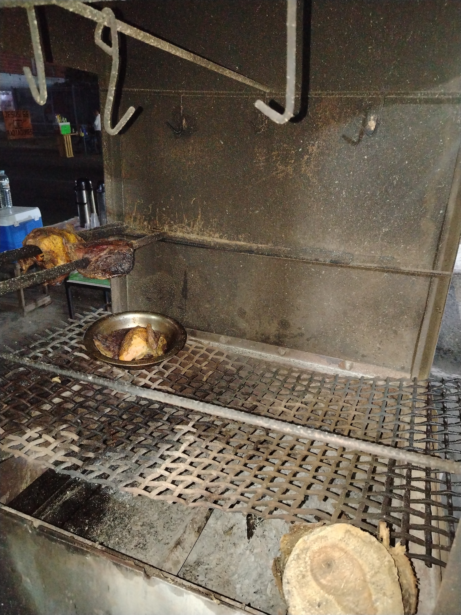 The image shows a food preparation area in Tinaquillo, Venezuela. A large metal grill sits within a metal enclosure, suggesting a custom-built or outdoor cooking setup. Above the grill, a rotisserie holds roasting meat. A metal bowl on the grill contains pieces of cooked meat. Below the grill is a pile of wood. The lighting is dim. Outside the immediate cooking area, there are glimpses of a cooler, thermoses, and a distant street scene.