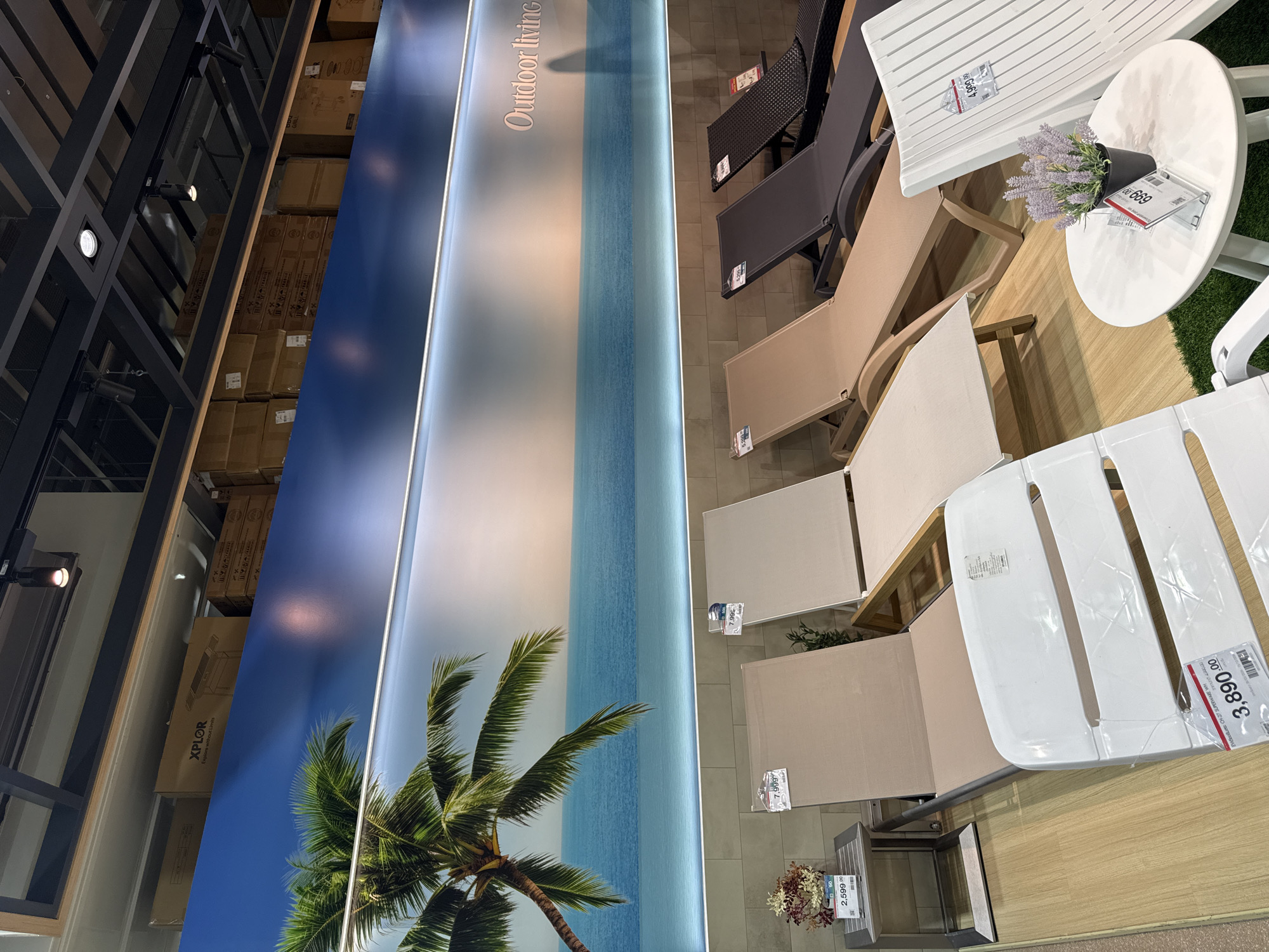 The image depicts an indoor display area, likely a furniture store, featuring several lounge chairs and a small side table. The background is a large mural of a tropical beach scene with a palm tree and ocean. The lounge chairs are of various colors, including beige, dark gray, and black, and are arranged in a row. A white plastic lounge chair is in the foreground, with a price tag attached. A small white round table with a potted lavender plant and a price tag is also visible. Several price tags are visible on the lounge chairs, indicating prices like 7,999.00, 2,599.00, and 3,890.00. The setting appears to be a retail environment in Baan Chaweng Noi, Thailand.