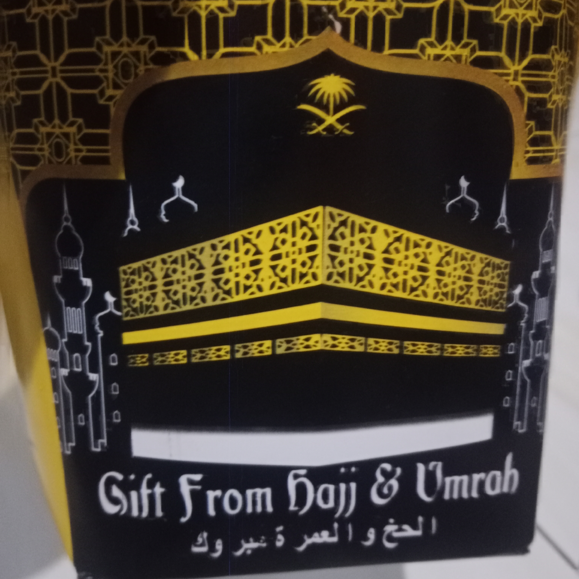 The image shows a close-up of a box, likely a gift box, with Islamic motifs. The box is predominantly black with gold and white accents. In the center, there is an illustration of the Kaaba in Mecca, depicted with a yellow patterned covering. Above the Kaaba, the Saudi Arabian emblem of a palm tree and two crossed swords is visible. On the left and right sides of the Kaaba illustration, there are stylized depictions of minarets. The box also features text in English, "Gift From Hajj & Umrah," and Arabic script below it. The background of the box has a geometric gold pattern. The object is placed on a white surface, possibly a table or shelf. The context of Tegowanu Kulon, Indonesia, is not directly inferable from the visual information in the image itself.