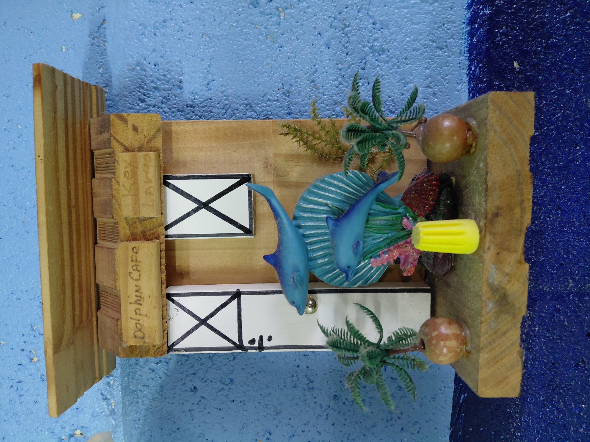 A wooden miniature diorama of the "Dolphin Cafe, Key Largo" is positioned on a blue surface. The diorama features two stylized blue dolphins, miniature palm trees, faux coral, and small wooden "windows" marked with black "X"s. The wooden frame displays "Dolphin Cafe" and "Key Largo" inscribed on its side. A yellow object is visible near the top right corner of the diorama. The background shows a textured blue surface. It is not possible to confirm the diorama's location in Cutler Bay based on visual information.