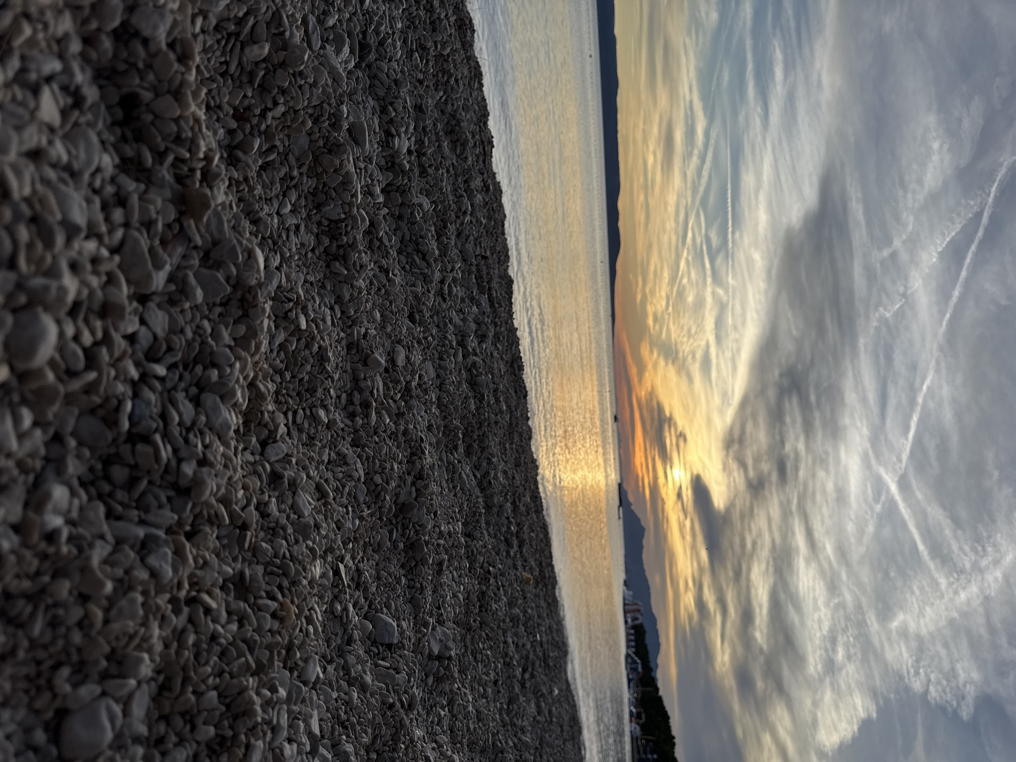 The image depicts a beach in Makarska, Croatia, during sunset. The foreground is dominated by a dense scattering of grey and white pebbles, characteristic of Makarska's beaches. Beyond the pebbles, a calm sea stretches towards the horizon, reflecting the warm hues of the setting sun. The sky above is a canvas of soft orange, yellow, and blue, streaked with wispy clouds and contrails, suggesting aerial activity. In the distance, the silhouette of a mountain range forms the backdrop, with a faint line of buildings and structures along the coastline. A few boats are visible on the water, and some inflatable water attractions are present near the shore, indicating a popular recreational area. The overall scene conveys a serene and picturesque evening ambiance.