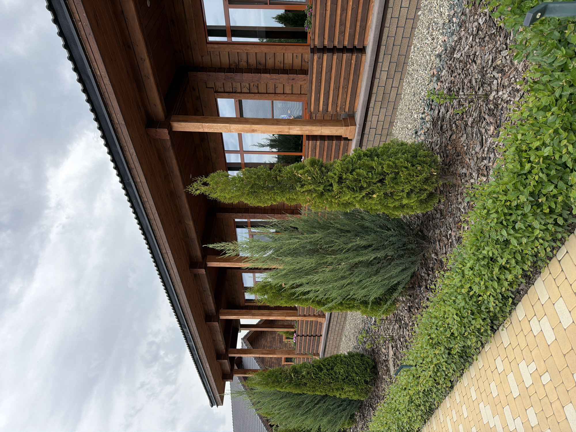 A low-angle shot of a wooden house with a porch in Kamyshin, Russia. The house is a light brown with a dark roof overhang. The porch is supported by wooden pillars that mirror the color of the house. There are large windows on the house, and a walkway made of rectangular tan and white stones leads to the entrance. Green bushes are planted along the walkway and in front of the house. The ground is covered with mulch. The sky is overcast.