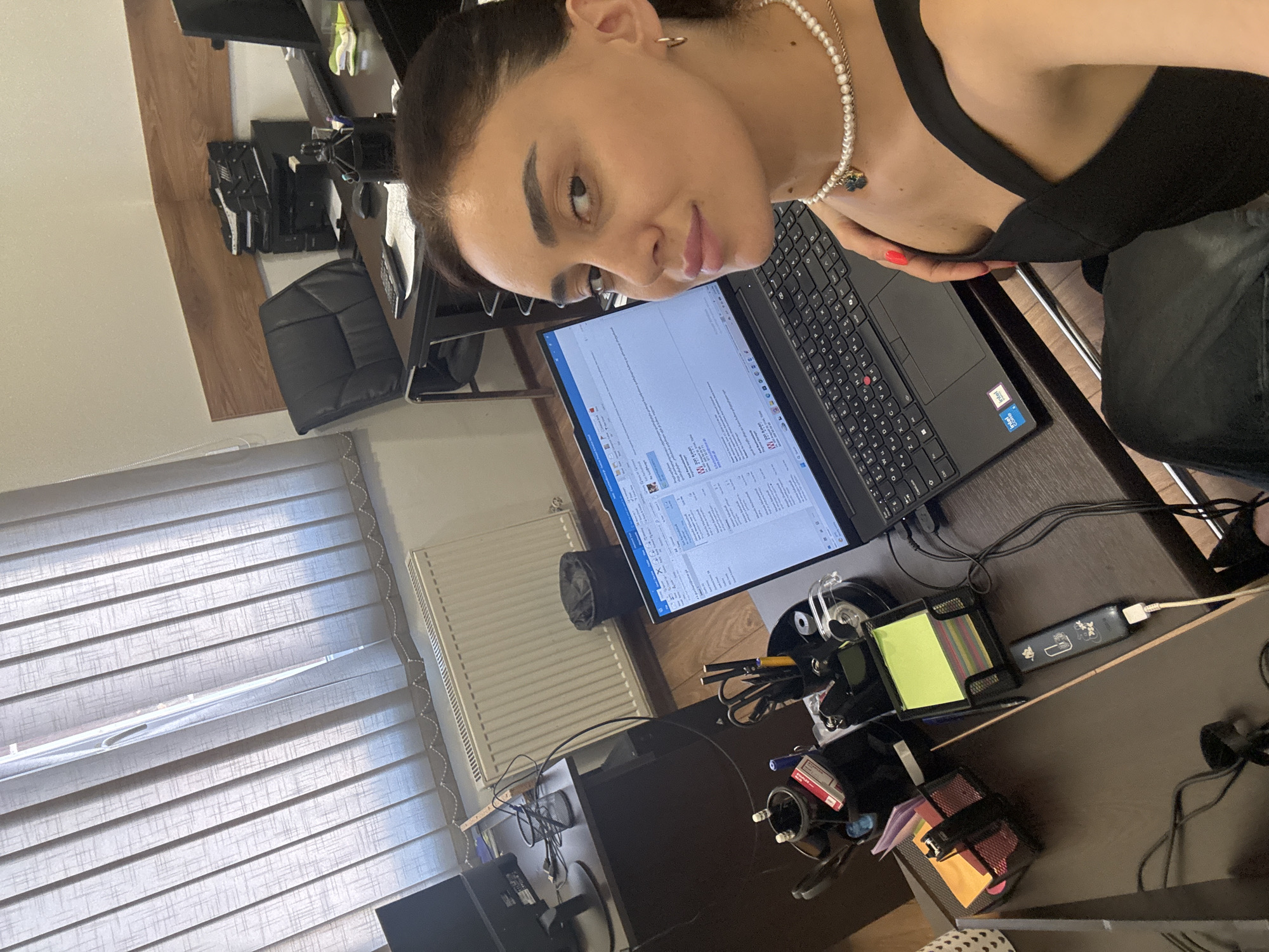 The image shows a woman taking a selfie in an office environment, likely in Tbilisi, Georgia. She is seated at a desk with a laptop in front of her. The laptop screen displays an email with text in Georgian. On the desk, there are various office supplies such as a stapler, pens, sticky notes, and a phone charger. The background reveals office furniture, including a desk with a printer and a black leather office chair. Vertical blinds cover a window, and a radiator is visible on the wall. The woman is wearing a black top and a pearl necklace.