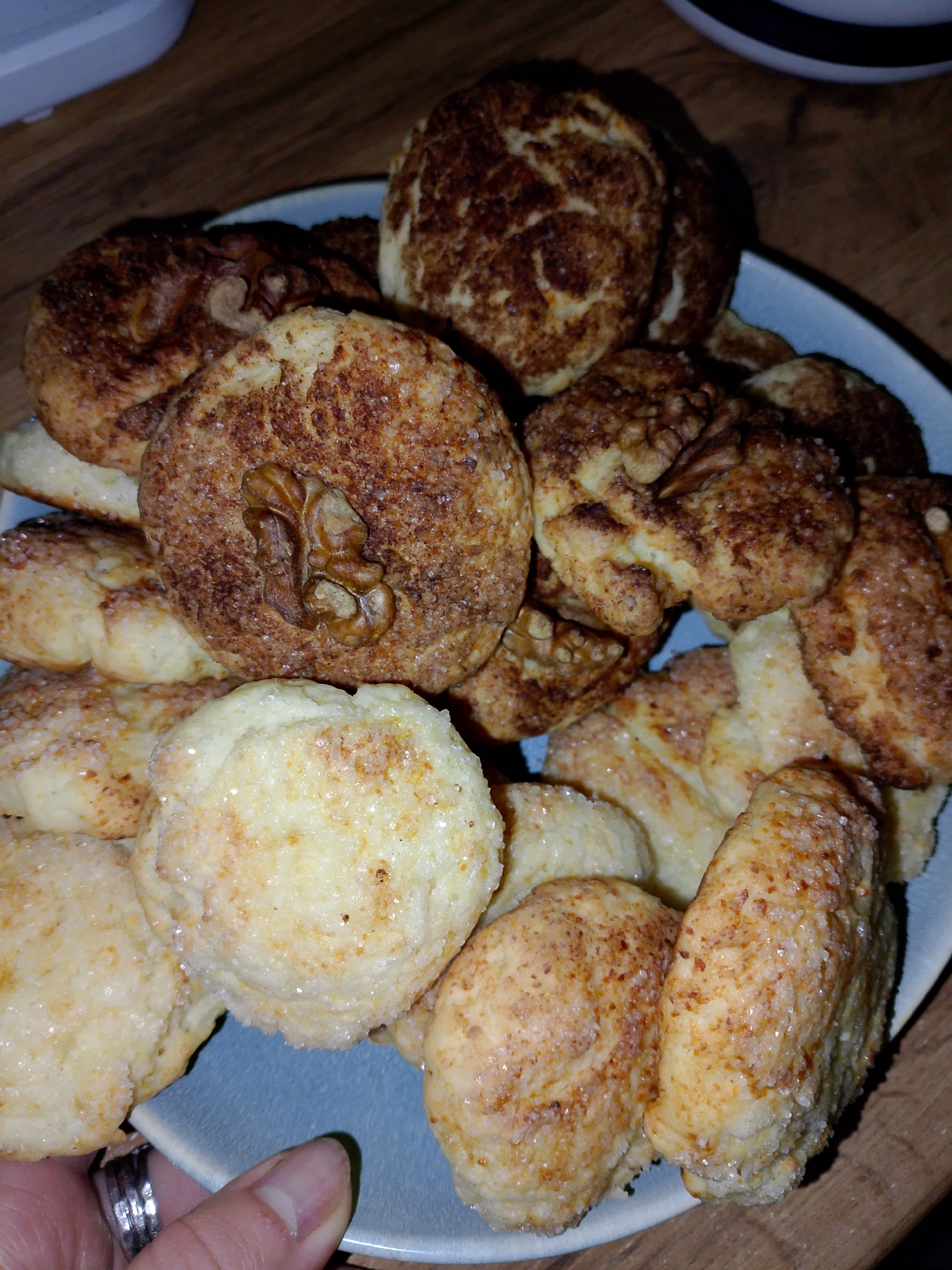 A plate of cookies is shown on a wooden surface. The plate contains several different types of cookies, some of which are decorated with a walnut. The cookies are golden brown in color and appear to be coated in sugar. A hand wearing a silver ring holds the plate. There is no indication of a specific location within Izhevsk, Russia, or any other geographic marker.