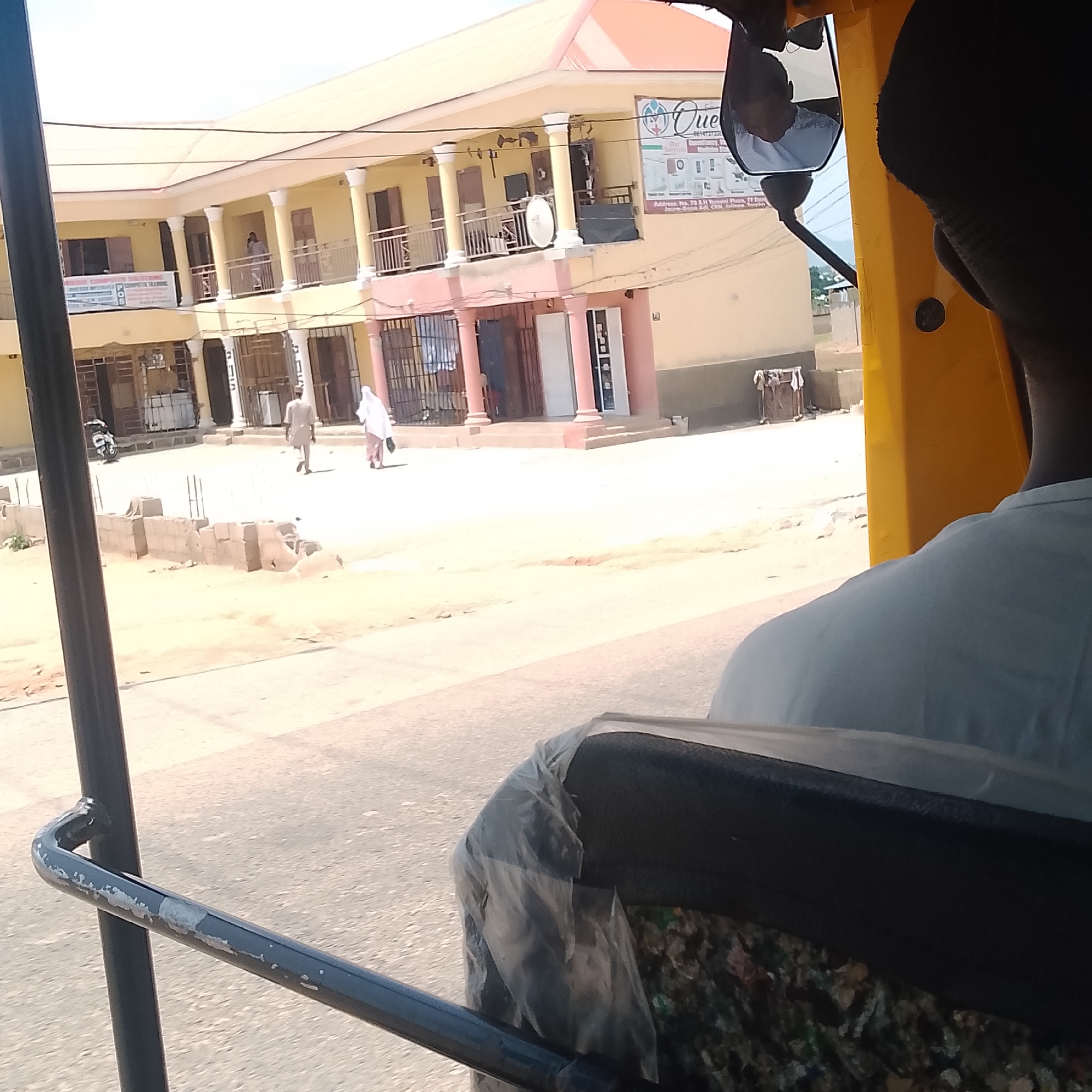 The image, taken from a three-wheeled vehicle, shows a street scene in Jalingo, Nigeria. In the background, a light yellow, two-story building with a pink trim is visible. The building has balconies with railings and classical-style columns. A banner advertising "QUEEN Computer Training" is displayed on the building. Two individuals are walking in front of the building: one man in a light-colored traditional outfit and a woman in a white hijab and dress. To the left of the building, a motorcycle is parked. In the foreground, part of the three-wheeled vehicle is visible, including a black metal bar and the back of a seat covered in plastic sheeting. The ground is dusty and uneven. The sky is overcast and the overall lighting suggests it is daytime.