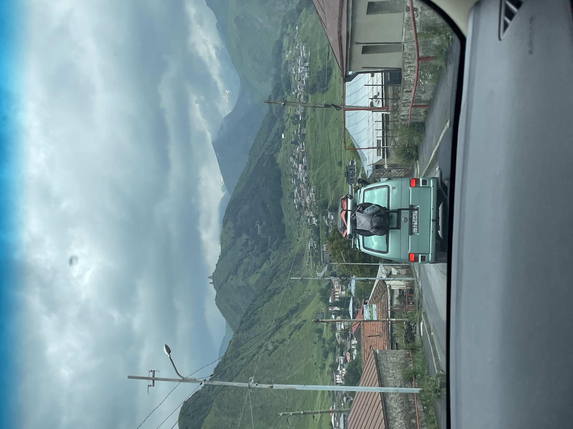 The image shows a view from inside a vehicle, looking forward through the windshield. A light blue van with luggage strapped to its roof is in front, driving on a road in a town. The town is nestled in a valley surrounded by lush green, mountainous terrain. Buildings with red-tiled roofs and stone walls are visible on either side of the road. In the background, a prominent church or monastery complex sits atop a hill, overlooking the town and the valley. The sky is overcast with dramatic clouds. The license plate of the van reads "321AHU1". The scene appears to be Stepantsminda, Georgia, given the distinctive landscape and the Gergeti Trinity Church visible on the hilltop. There are no people directly visible in the scene, besides implied occupants of the van.
