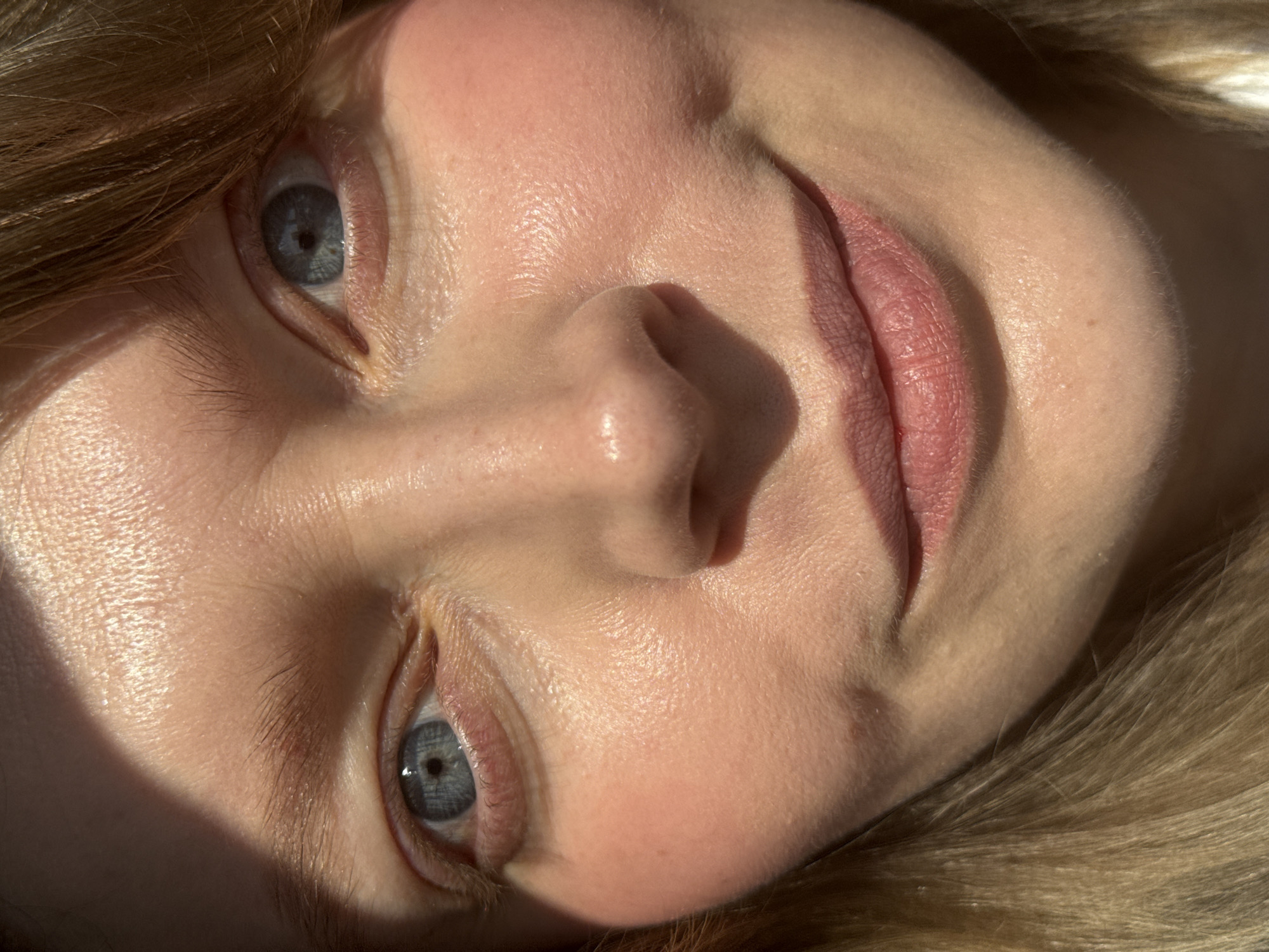 A close-up image features the face of a fair-skinned person with light blonde hair, positioned in a tilted manner relative to the viewer. The person has light blue eyes and pink lips. The lighting suggests a natural outdoor setting. No city or location is discernible from the image.